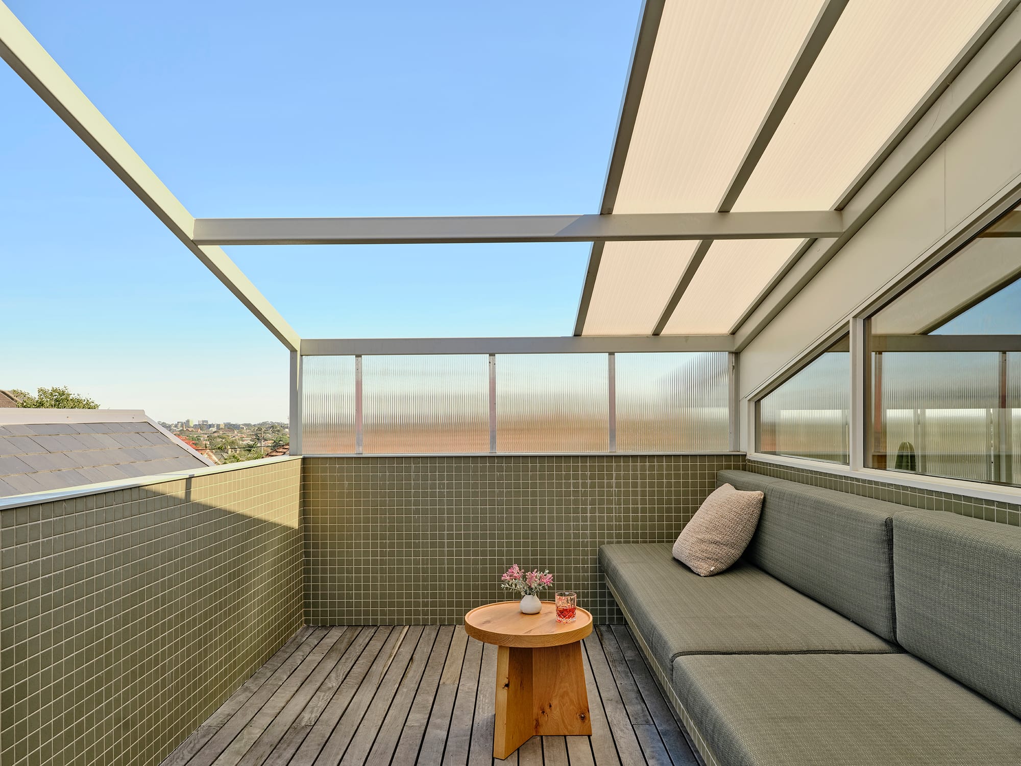 Salmon House by True Story. Photography by Dean Bradley.A small rooftop terrace features olive-toned tiled walls, a cushioned bench, and a timber coffee table, partially shaded by a translucent roof.
