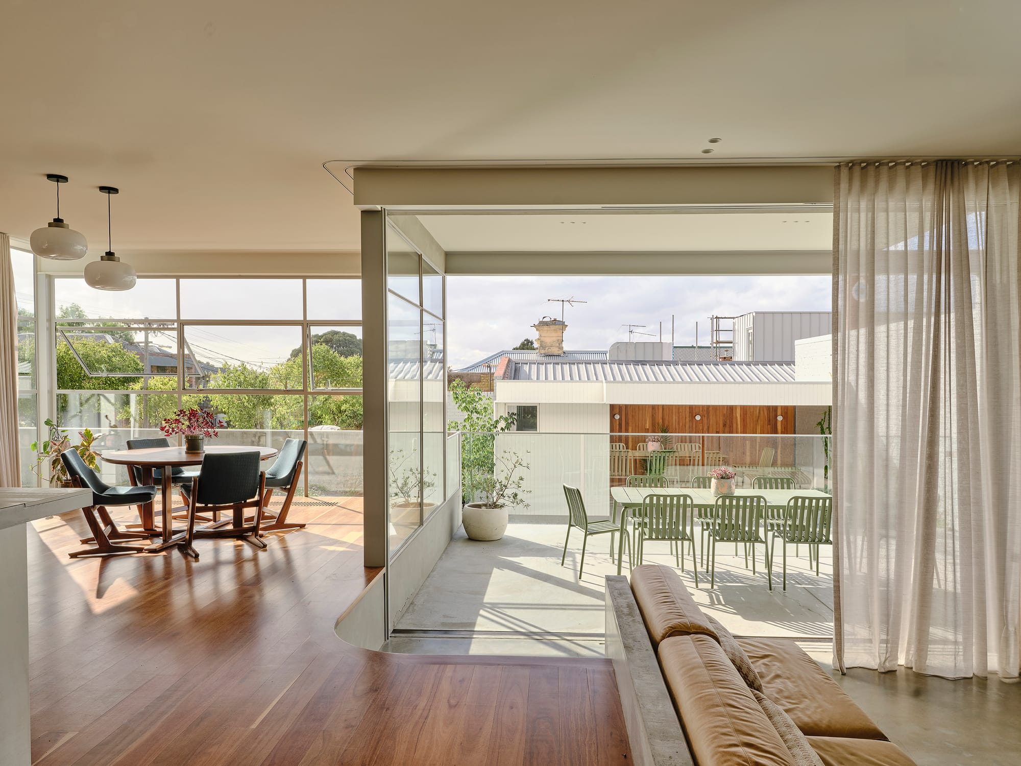 Salmon House by True Story. Photography by Dean Bradley.Open-plan living and dining area with timber floors, soft curtains, and a seamless connection to an outdoor terrace furnished with green chairs and potted plants.