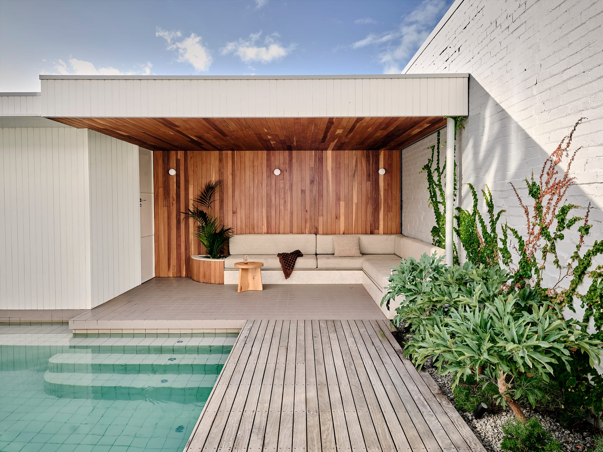 Salmon House by True Story. Photography by Dean Bradley.Poolside pavilion with timber ceiling and wall panelling, built-in cream seating, and lush planting along a white brick wall.