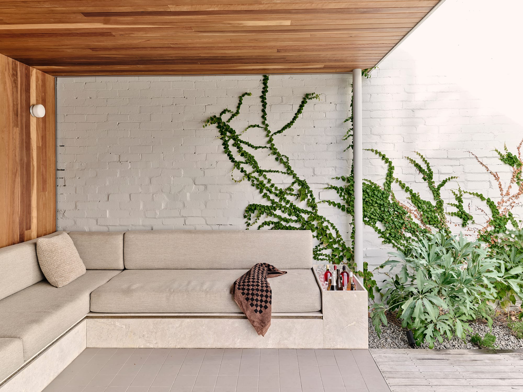 Salmon House by True Story. Photography by Dean Bradley. Outdoor seating nook with cream cushioned benches, timber ceiling, trailing vines on a white brick wall, and integrated drinks storage.