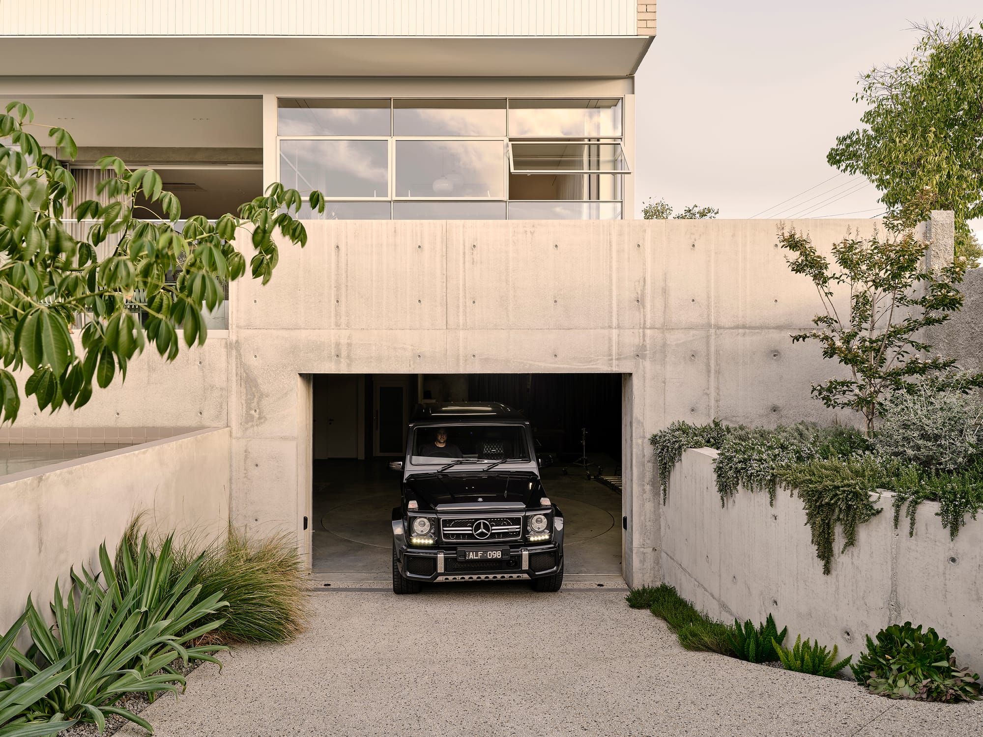 Salmon House by True Story. Photography by Dean Bradley.A black Mercedes G-Wagon emerges from a concrete garage beneath a modern home, framed by lush landscaping and minimalist architecture. 