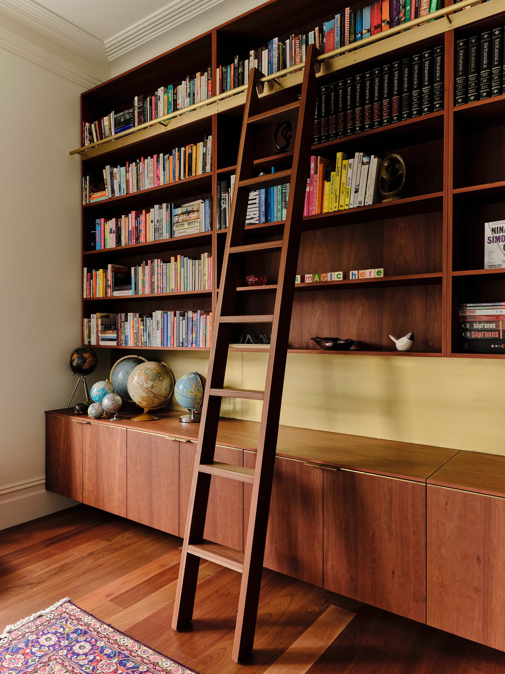 Salmon House by True Story. Photography by Dean Bradley.Tall timber bookshelf with globes and a library ladder against warm wood cabinetry.