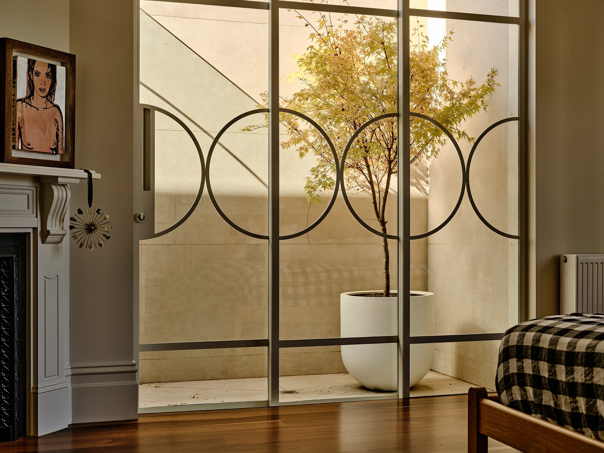 Salmon House by True Story. Photography by Dean Bradley. Sunlit courtyard tree in a white planter viewed through decorative glass doors from a bedroom with timber flooring and a fireplace.
