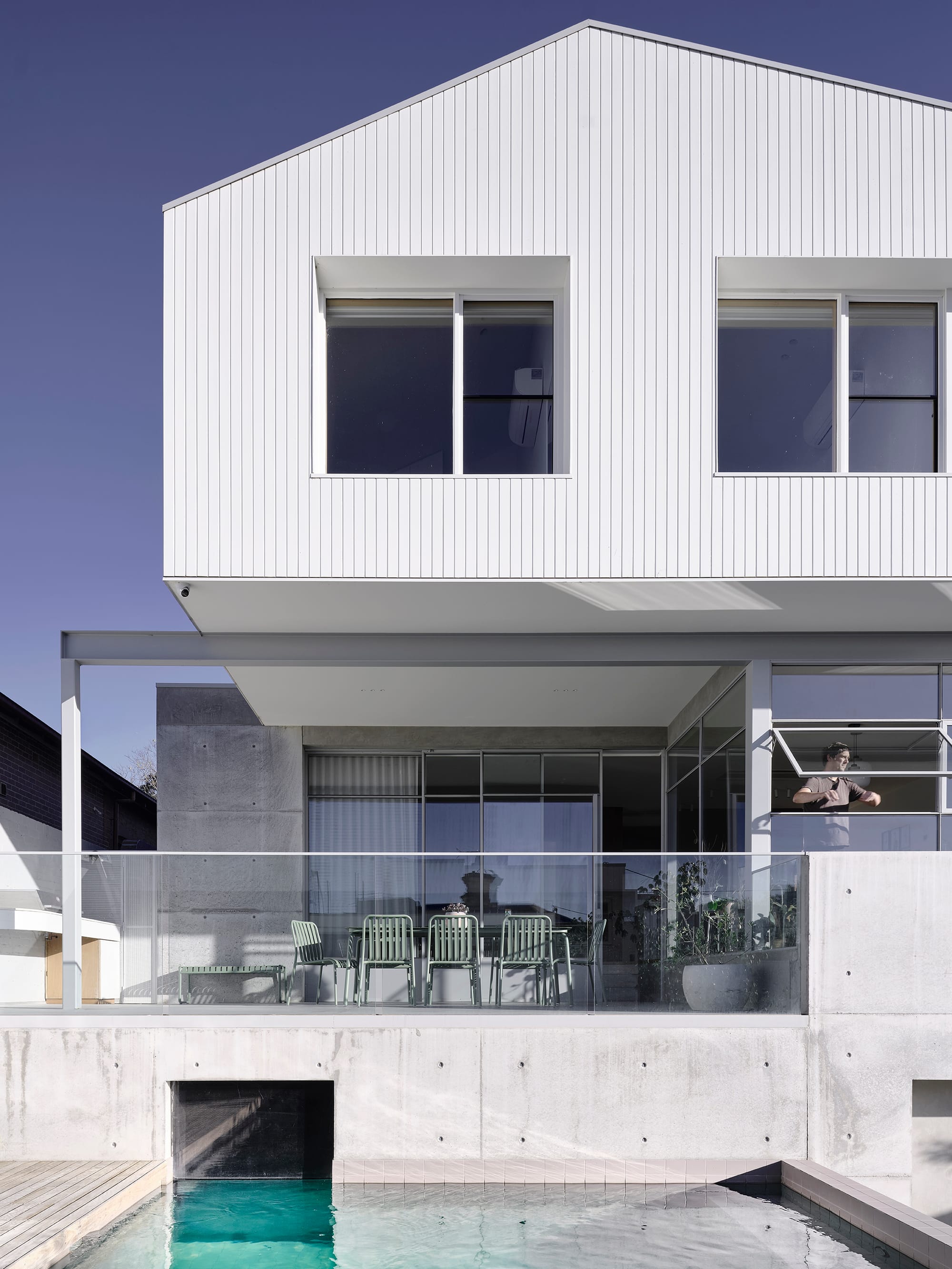 Salmon House by True Story. Photography by Dean Bradley. White gable-roofed home with a concrete base and glass balustrade, overlooking a turquoise pool and outdoor dining area.