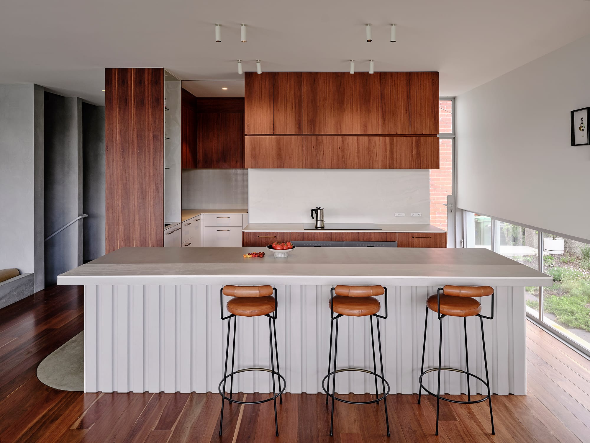 Salmon House by True Story. Photography by Dean Bradley.Modern kitchen with timber cabinetry, a concrete island featuring fluted detailing, and three leather-topped bar stools.
