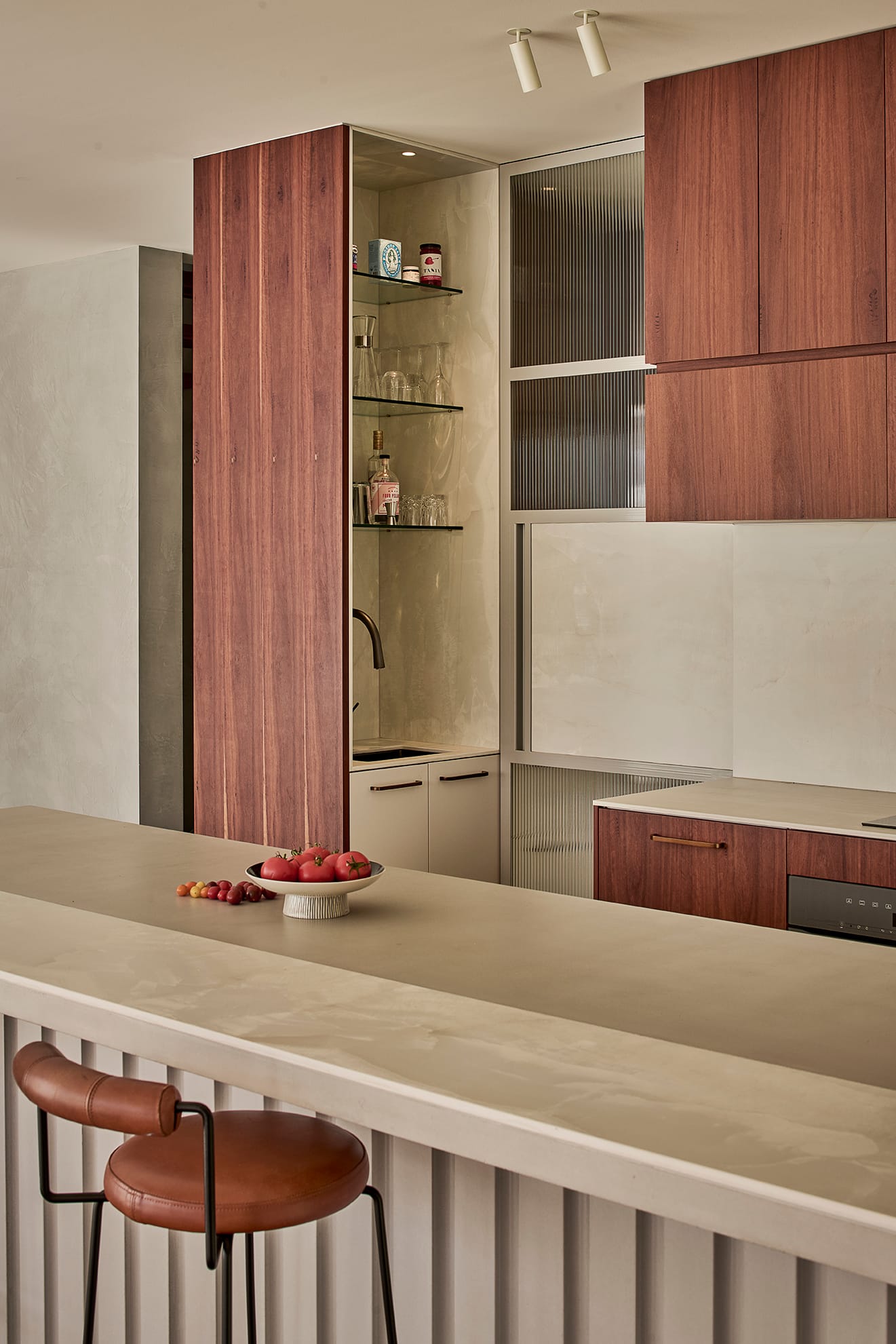 Salmon House by True Story. Photography by Dean Bradley. Detail view of a modern kitchen with timber cabinetry, open bar shelving, integrated sink, and fluted kitchen island with leather barstool.