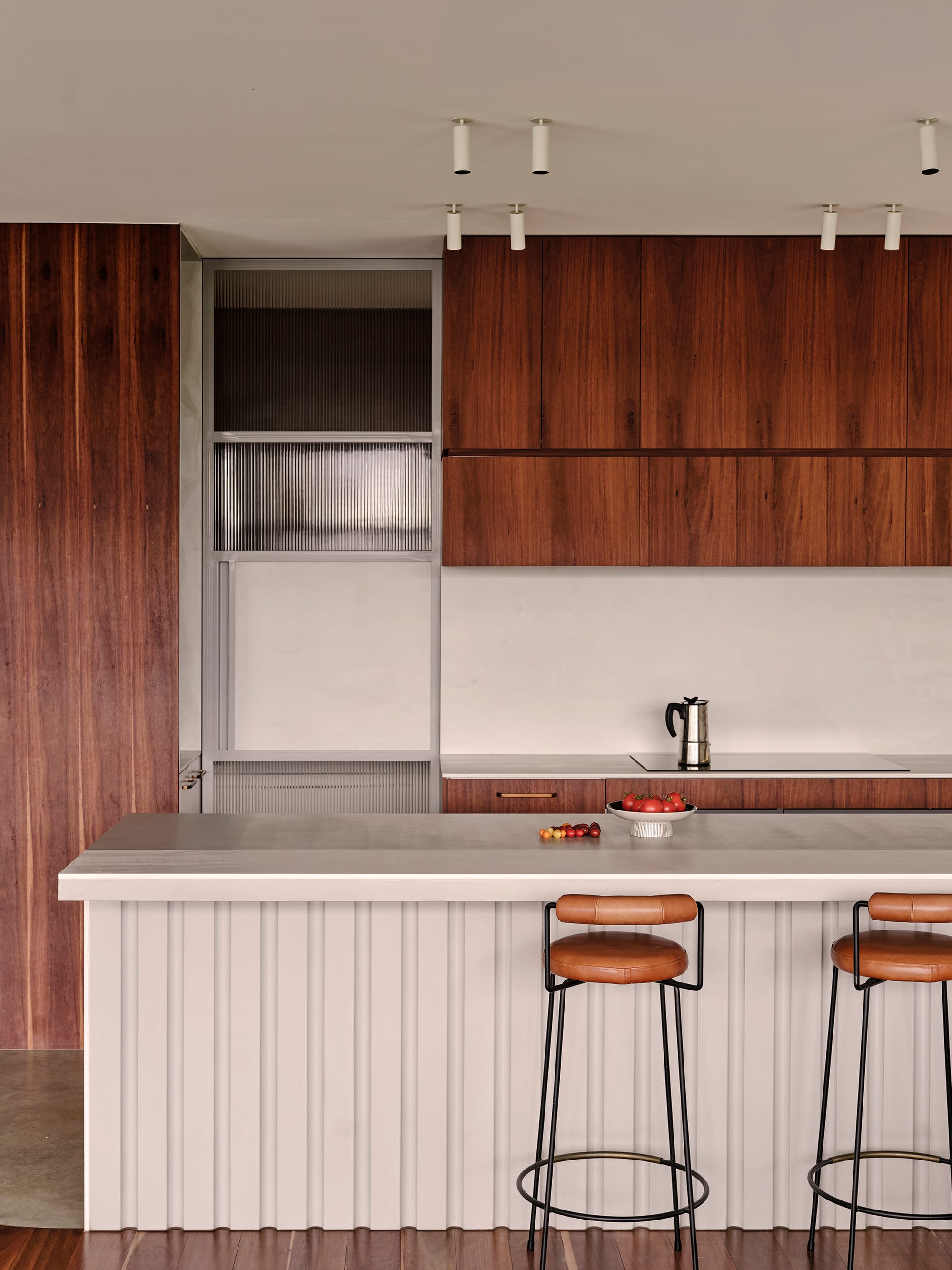 Salmon House by True Story. Photography by Dean Bradley.Detail view of a modern kitchen with timber cabinetry, open bar shelving, integrated sink, and fluted kitchen island with leather barstool.