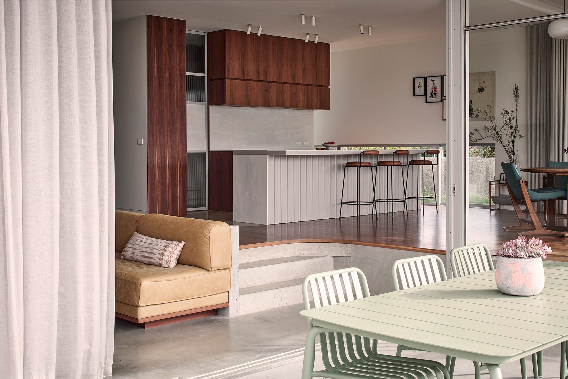 Salmon House by True Story. Photography by Dean Bradley.View from outdoor dining area into an open-plan kitchen and living space, featuring timber cabinetry, bar stools, and a tan leather built-in sofa.