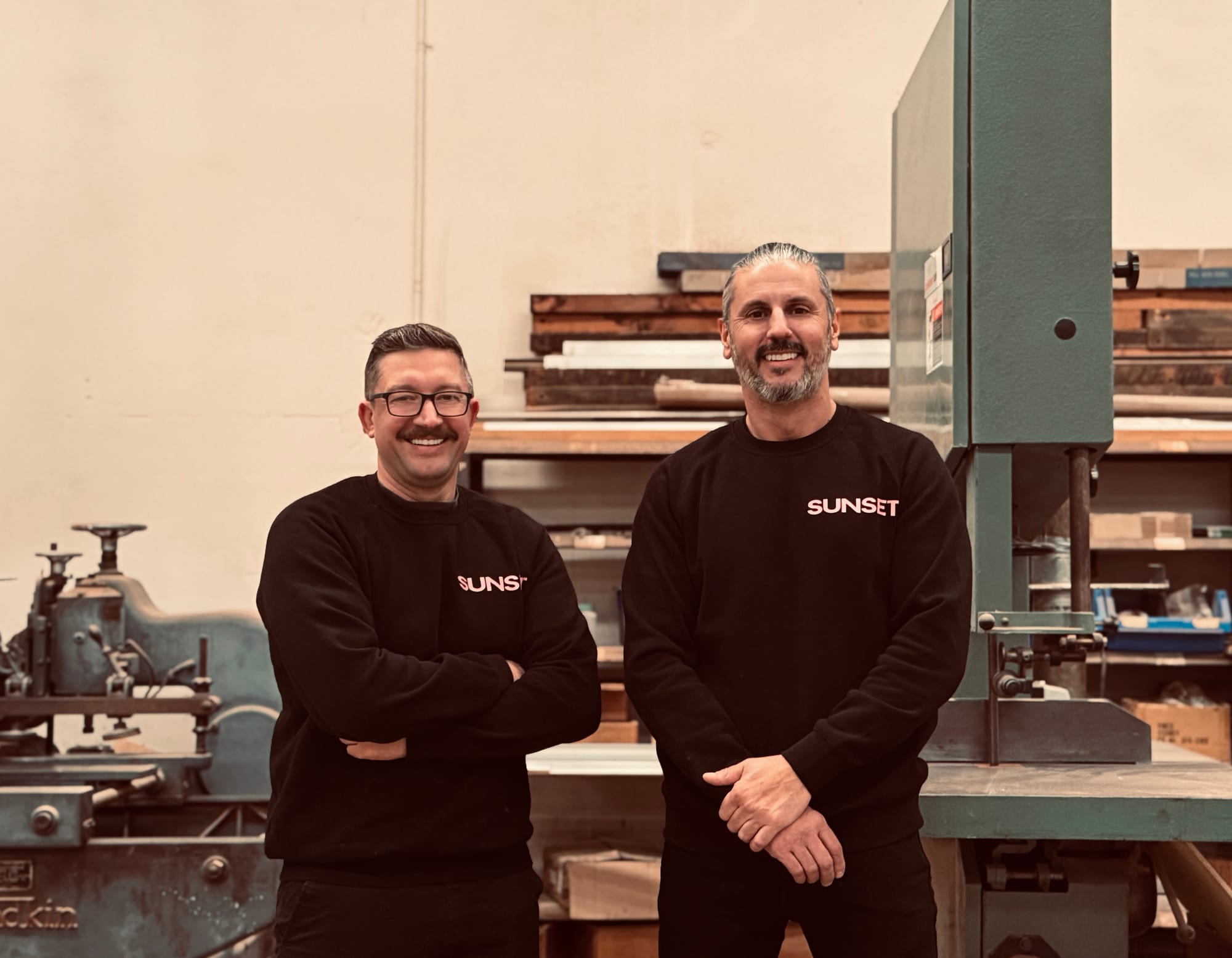 Sunset Joinery. Photography by Sunset Joinery. Two men wearing matching SUNSET jumpers stand smiling in a workshop, surrounded by timber, tools, and industrial equipment.
