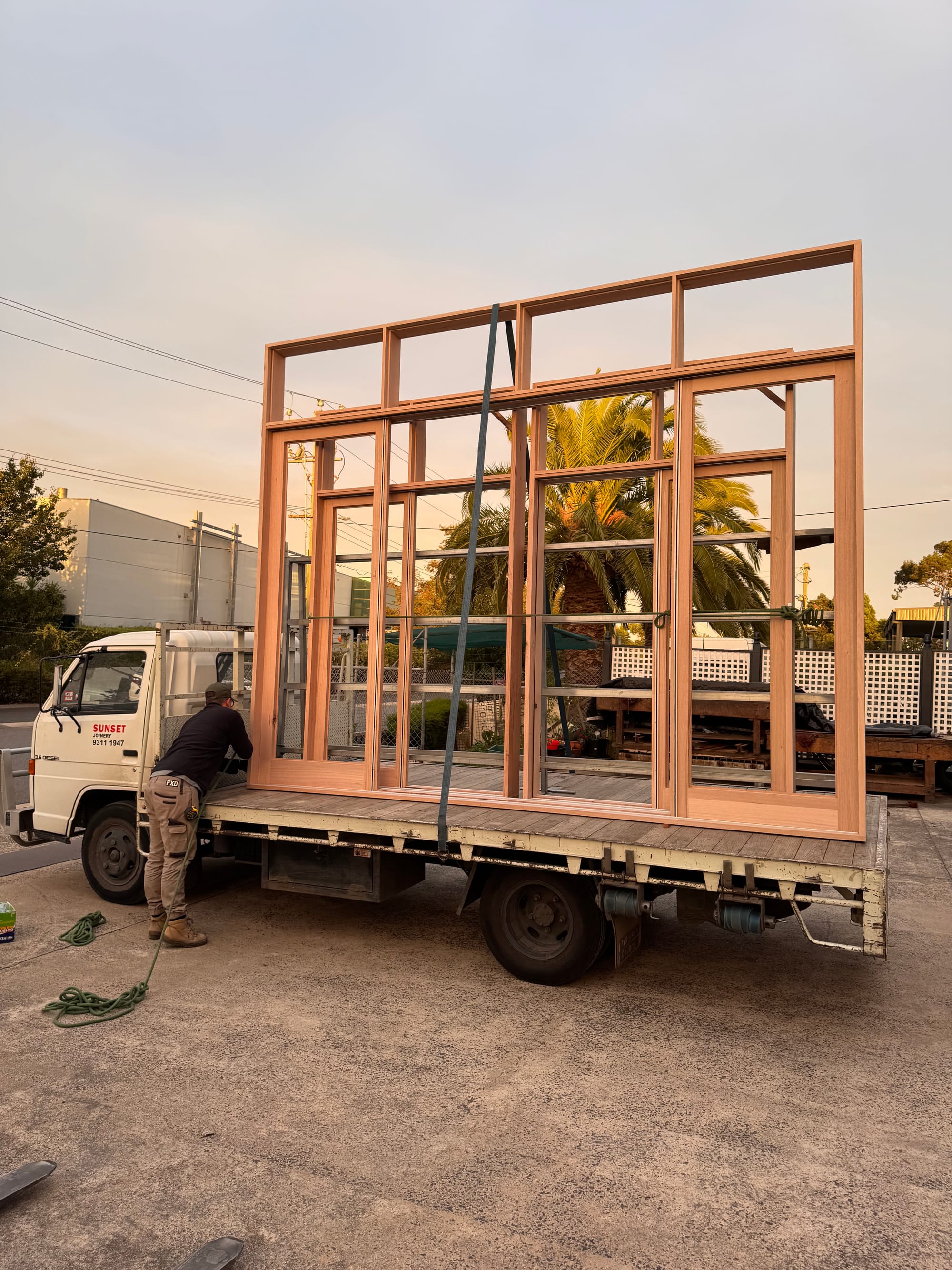 Sunset Joinery. Photography by Sunset Joinery.Worker securing a large custom timber window frame onto a flatbed truck at sunset, with a “SUNSET” branded vehicle in the background.