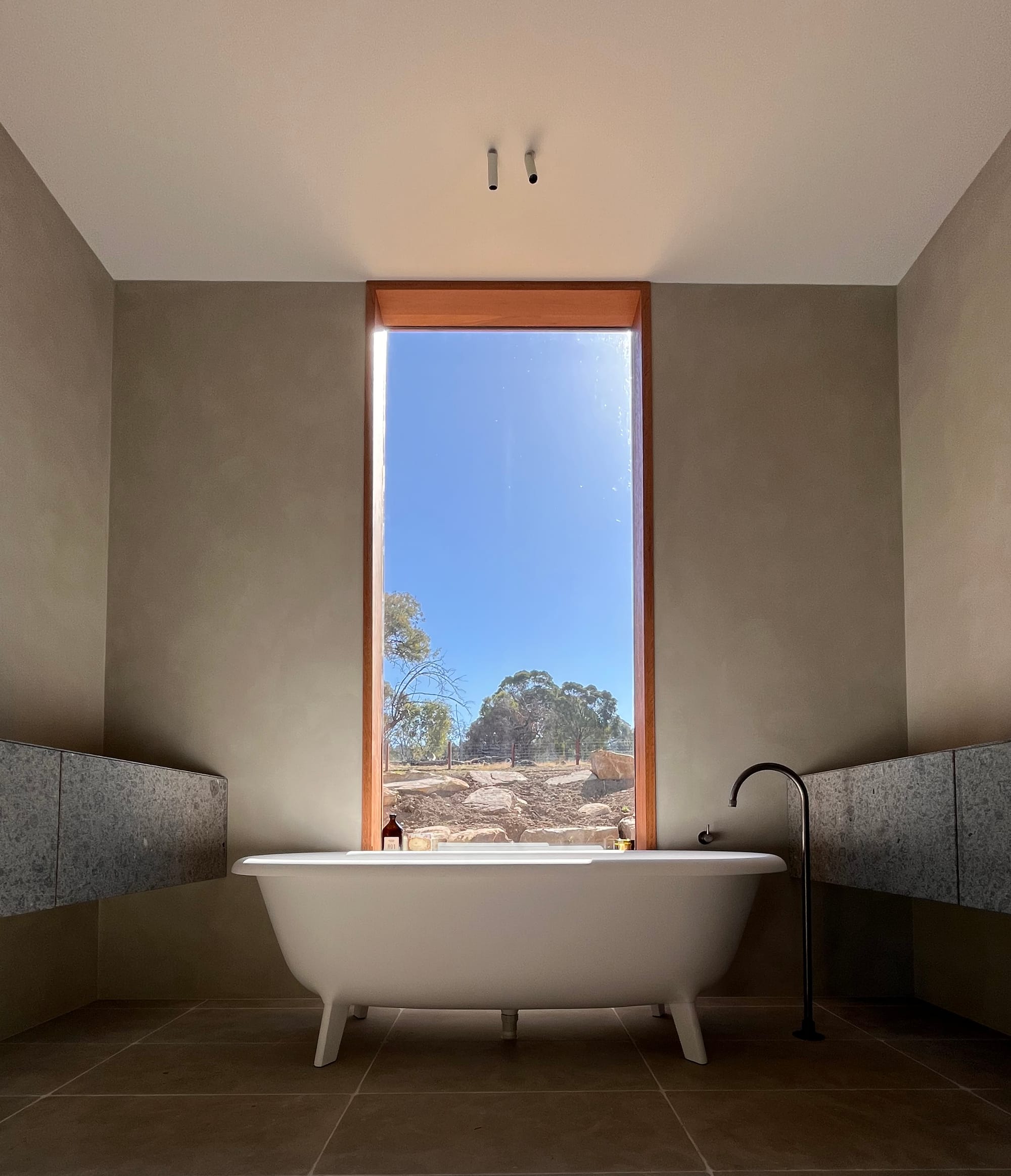 Sunset Joinery. Photography by Sunset Joinery.Minimalist bathroom with a freestanding white tub centered beneath a tall picture window, showcasing a natural landscape view and framed by smooth grey walls and stone shelving.