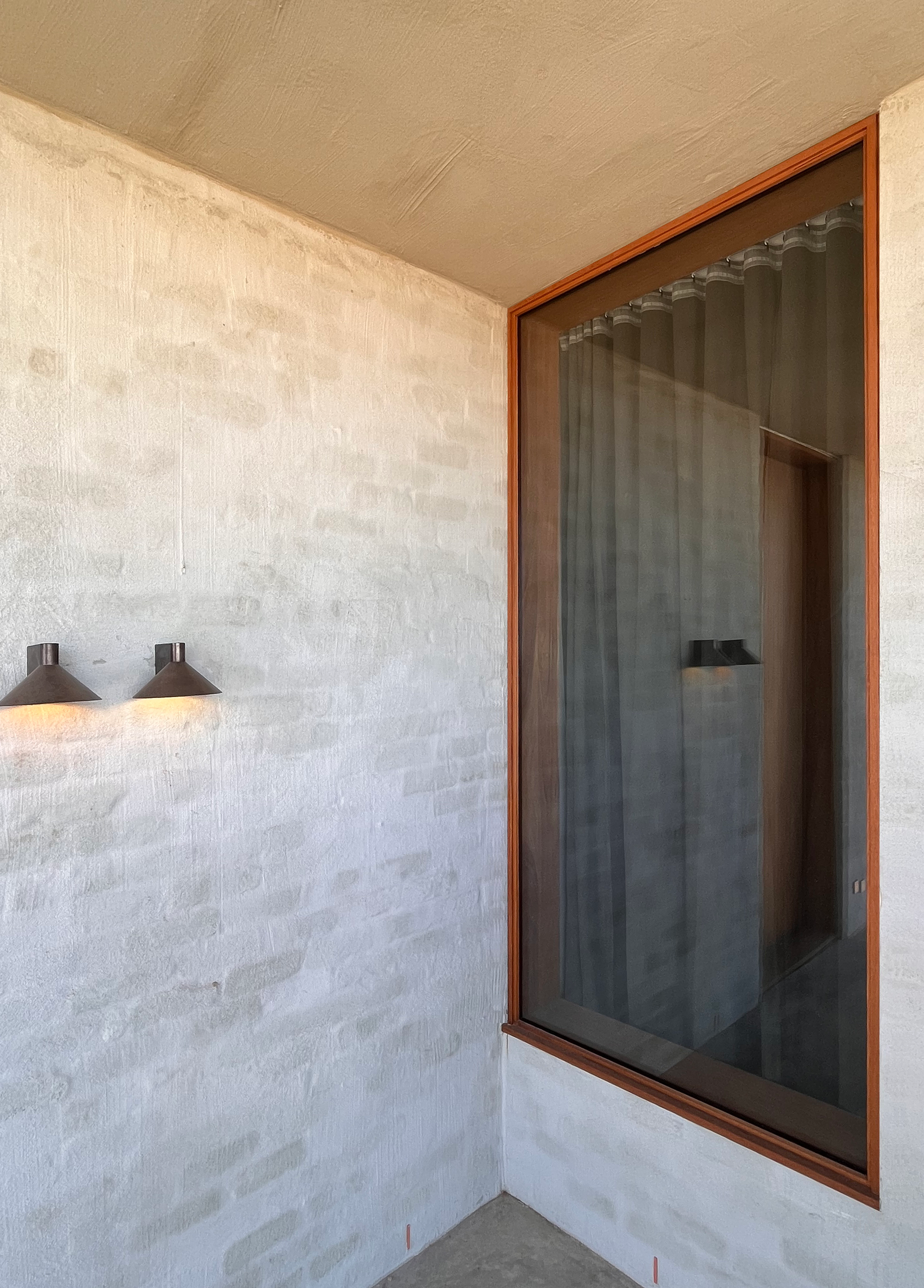 Sunset Joinery. Photography by Sunset Joinery.Minimal corner detail with whitewashed brick walls, a timber-framed window with sheer curtains, and two sculptural black wall lights casting a warm glow.
