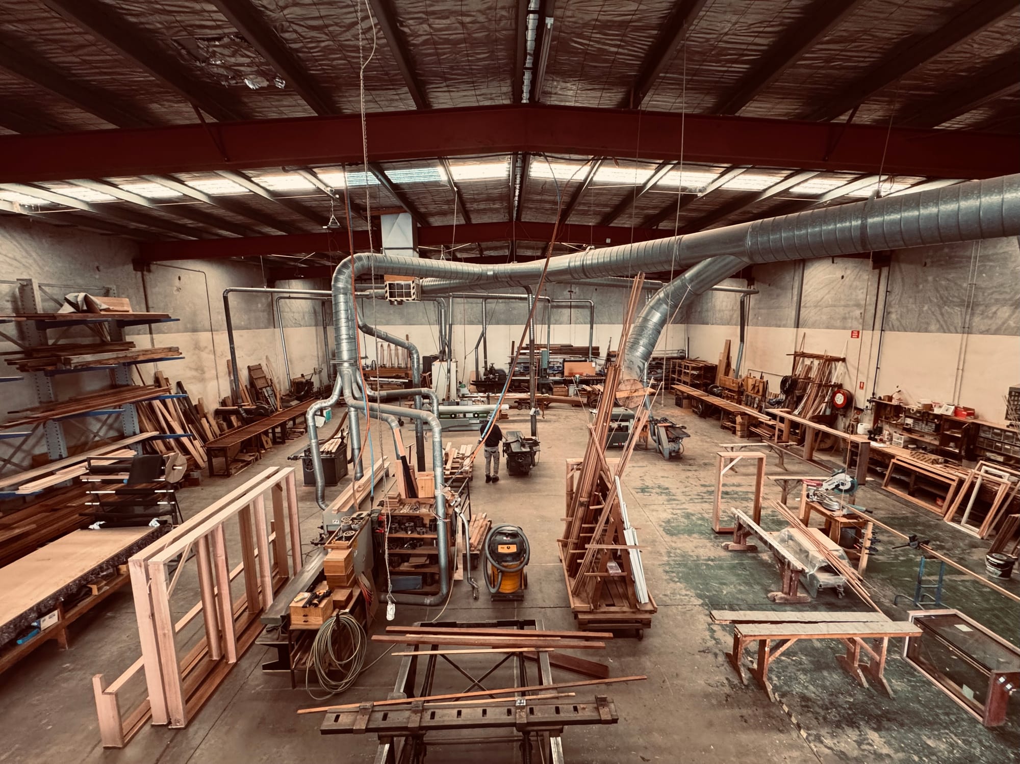 Sunset Joinery. Photography by Sunset Joinery.Wide view of a timber workshop with industrial ventilation, workbenches, machinery, and stacks of timber and joinery pieces in progress.