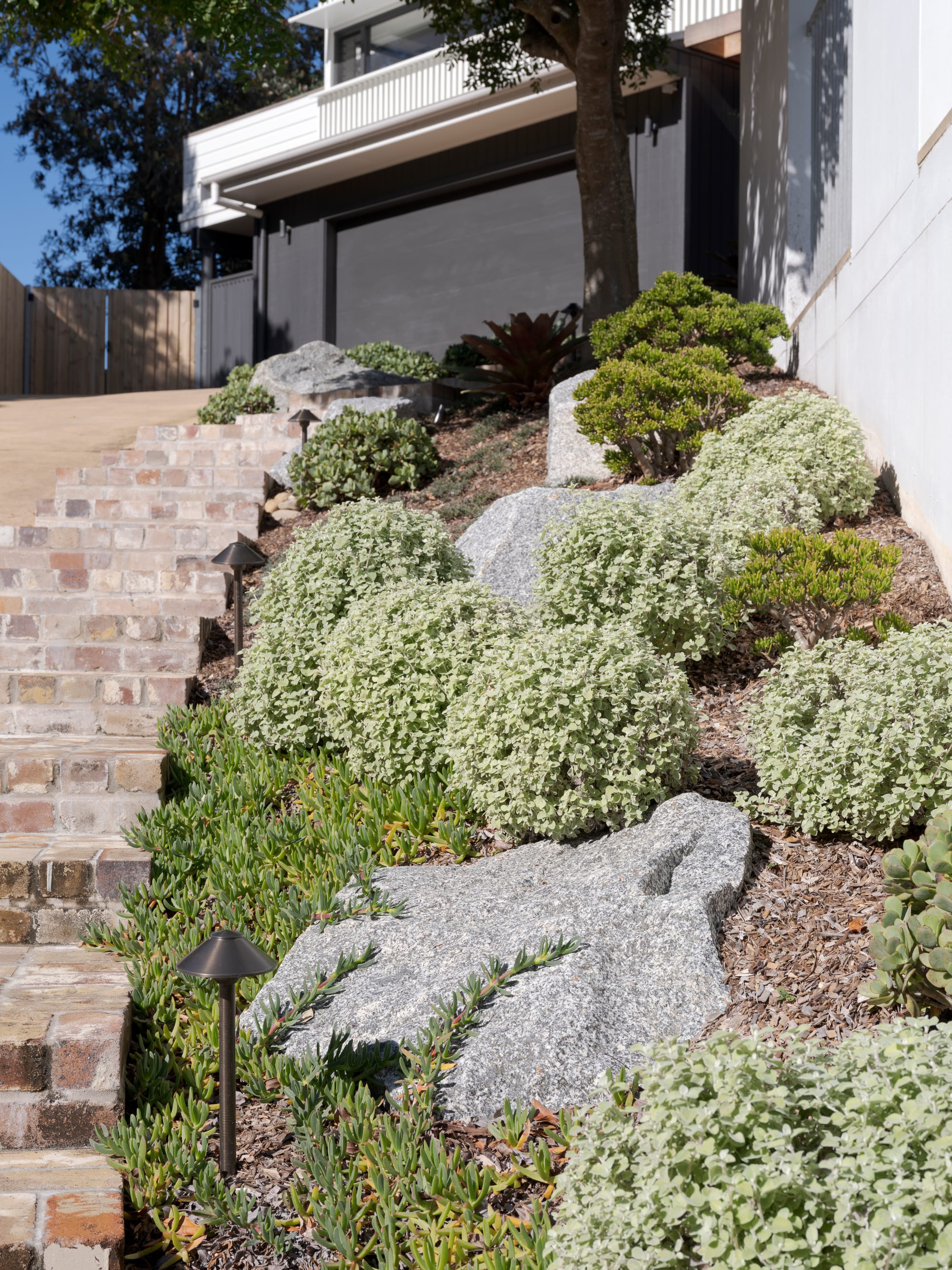Three Points House by BIJL Architecture. Photography by Tom Ferguson.Tiered front garden with brick steps, sculptural boulders, drought-tolerant plants, and layered greenery leading up to a contemporary home with dark garage doors.