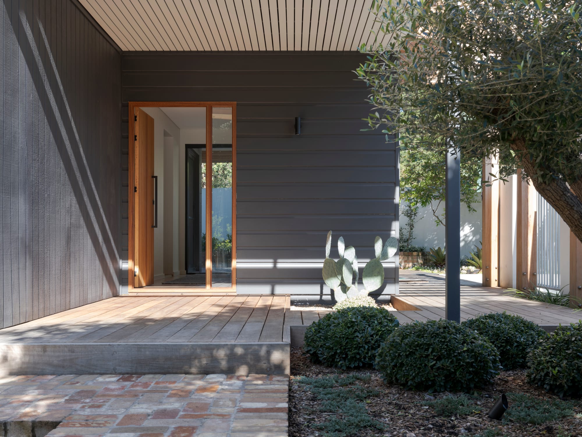 Three Points House by BIJL Architecture. Photography by Tom Ferguson.Modern entry porch with dark horizontal cladding, timber decking, glass-framed front door, and soft landscaping including a sculptural cactus and manicured shrubs.