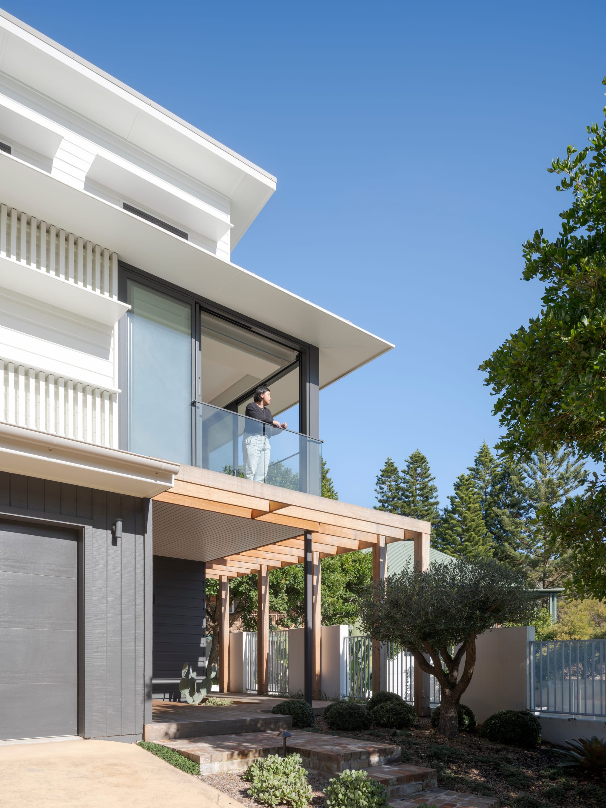 Three Points House by BIJL Architecture. Photography by Tom Ferguson.Modern entryway with dark horizontal cladding, timber decking, glass-framed front door, and minimalist landscaping including a sculptural cactus and manicured shrubs.Modern two-storey home with a mix of white and dark cladding, glass balcony, timber pergola, and landscaped front garden, viewed under a clear blue sky.