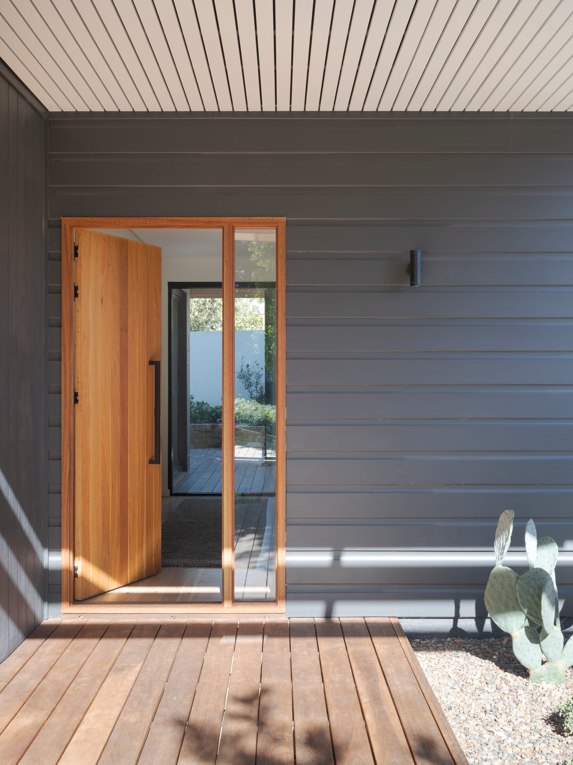 Three Points House by BIJL Architecture. Photography by Tom Ferguson.Timber front door with glass side panel set into a dark horizontal-clad facade, opening onto a warm-toned timber deck with minimalist desert-style landscaping.