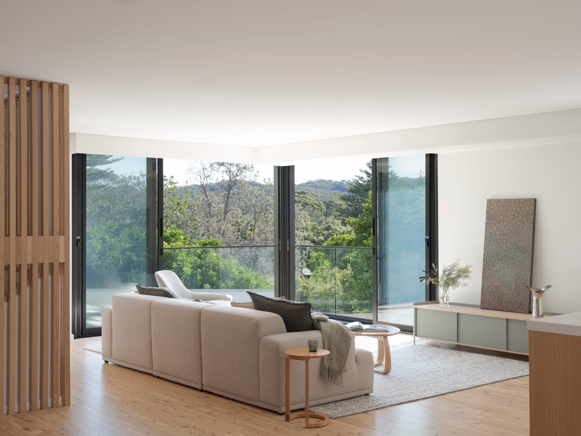 Three Points House by BIJL Architecture. Photography by Tom Ferguson.Living area with large corner glazing and forest views, light timber floors, neutral-toned sofa, and a minimal sideboard with artwork.