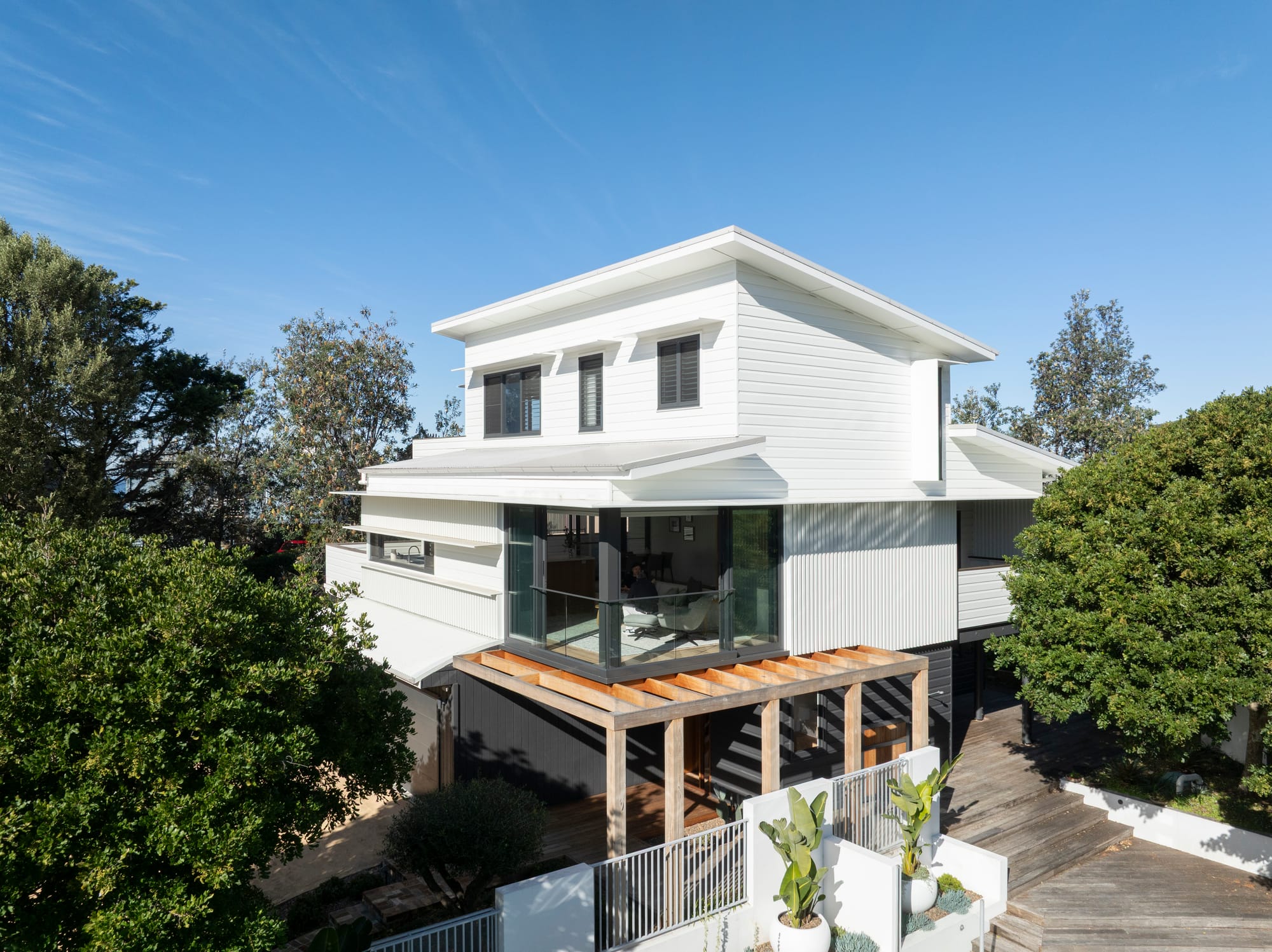 Three Points House by BIJL Architecture. Photography by Tom Ferguson.Modern two-storey home with a white weatherboard and corrugated exterior, corner glass window, timber pergola, and landscaped entry framed by mature trees and tiered planters.