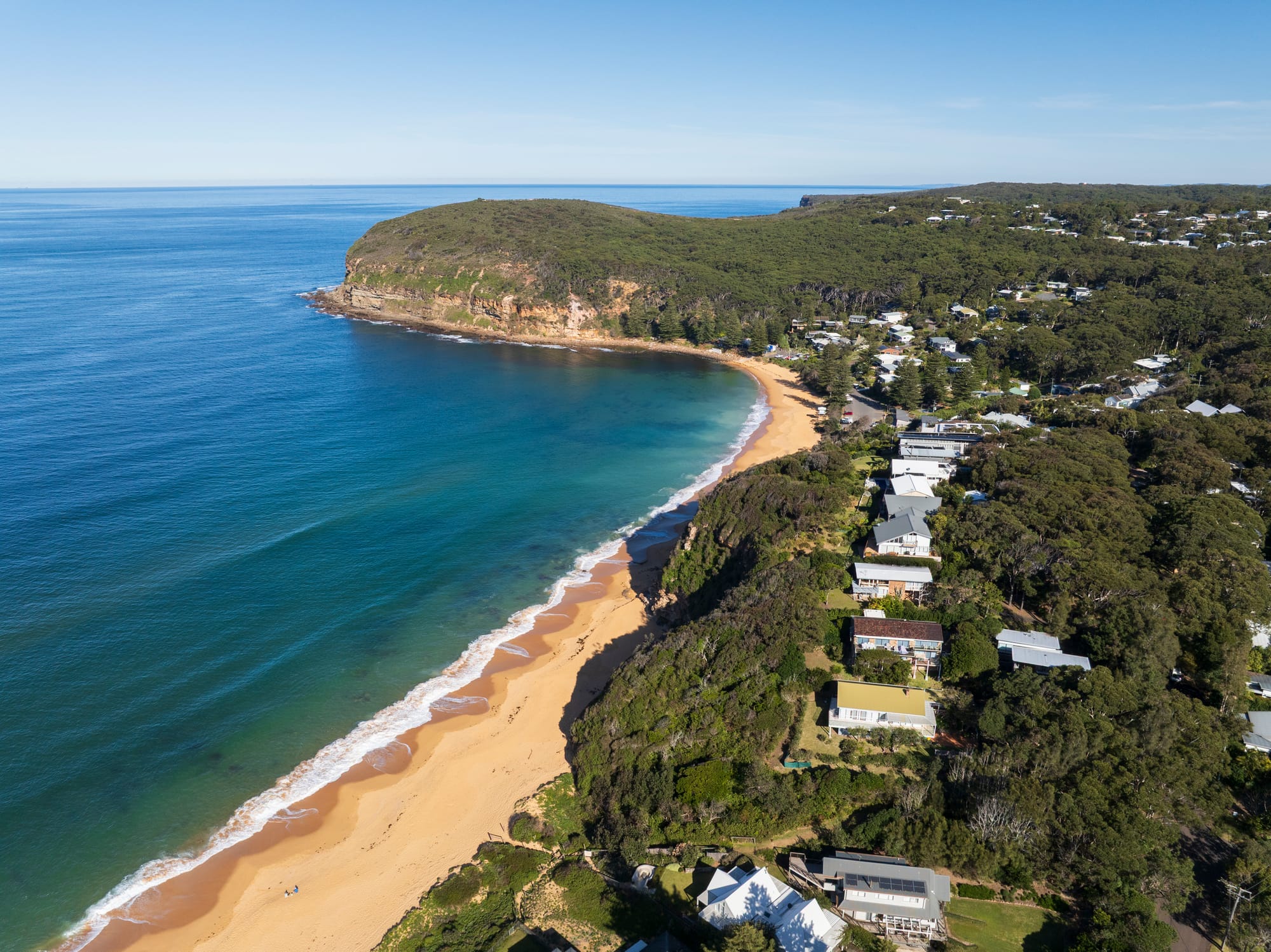 Three Points House by BIJL Architecture. Photography by Tom Ferguson.Aerial view of a secluded beach with golden sand, clear blue water, forested headland, and houses nestled along the coastline.