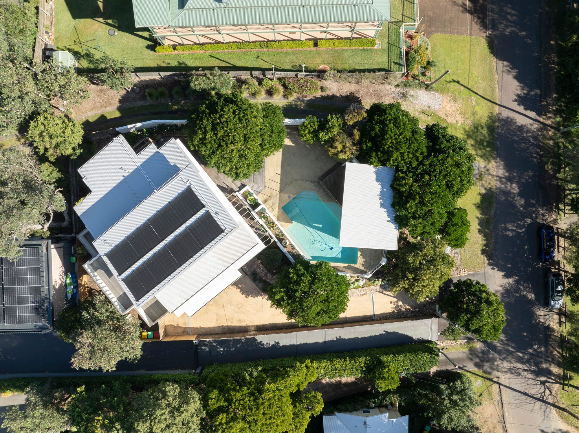 Three Points House by BIJL Architecture. Photography by Tom Ferguson.Aerial view of a residential property featuring a white house with solar panels, landscaped yard, swimming pool, and poolside pavilion surrounded by trees and neighboring houses.