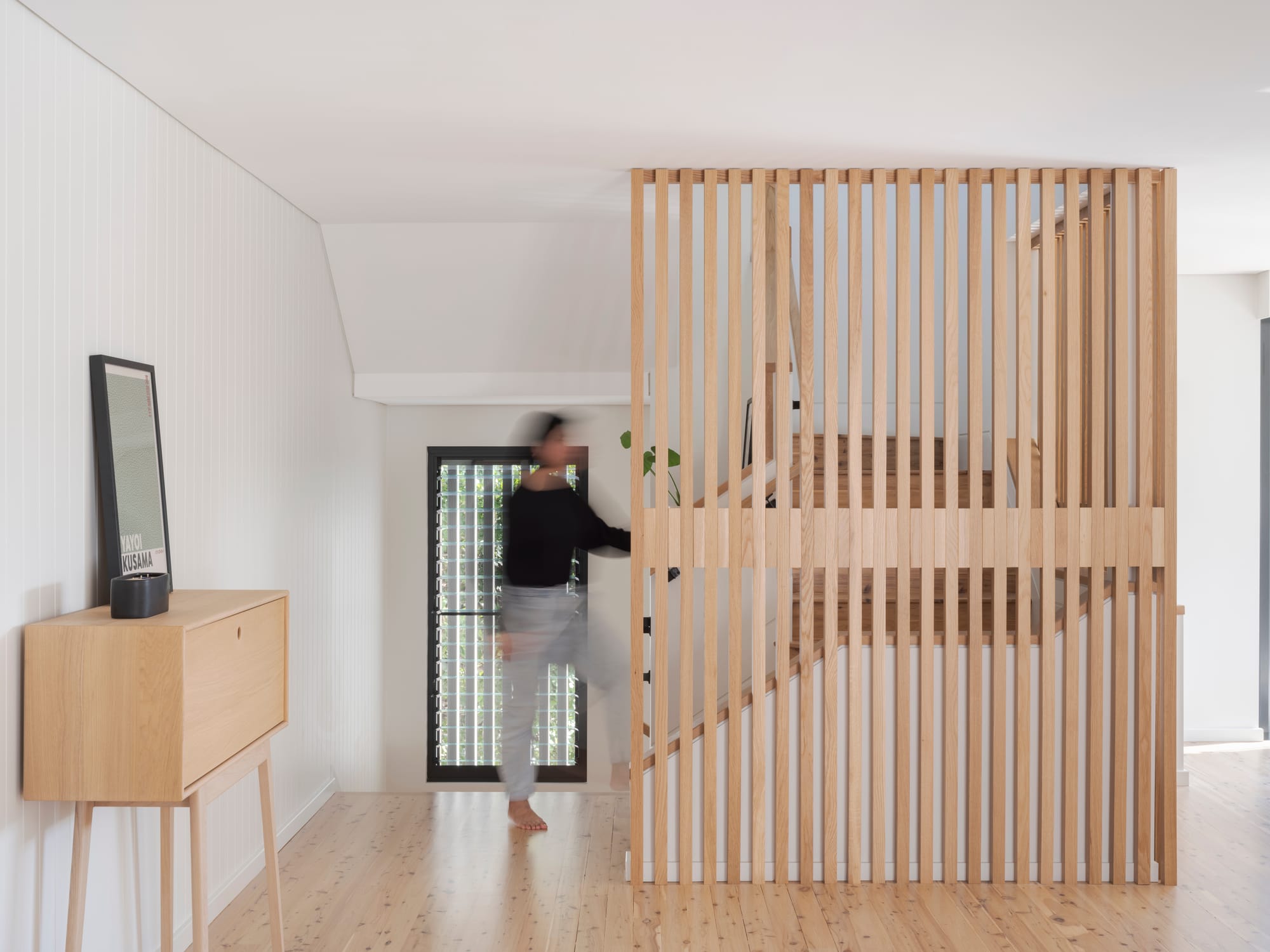 Three Points House by BIJL Architecture. Photography by Tom Ferguson.Person walking past light timber batten screen enclosing staircase in bright hallway with pale flooring, white walls, and a framed poster atop a timber cabinet. 