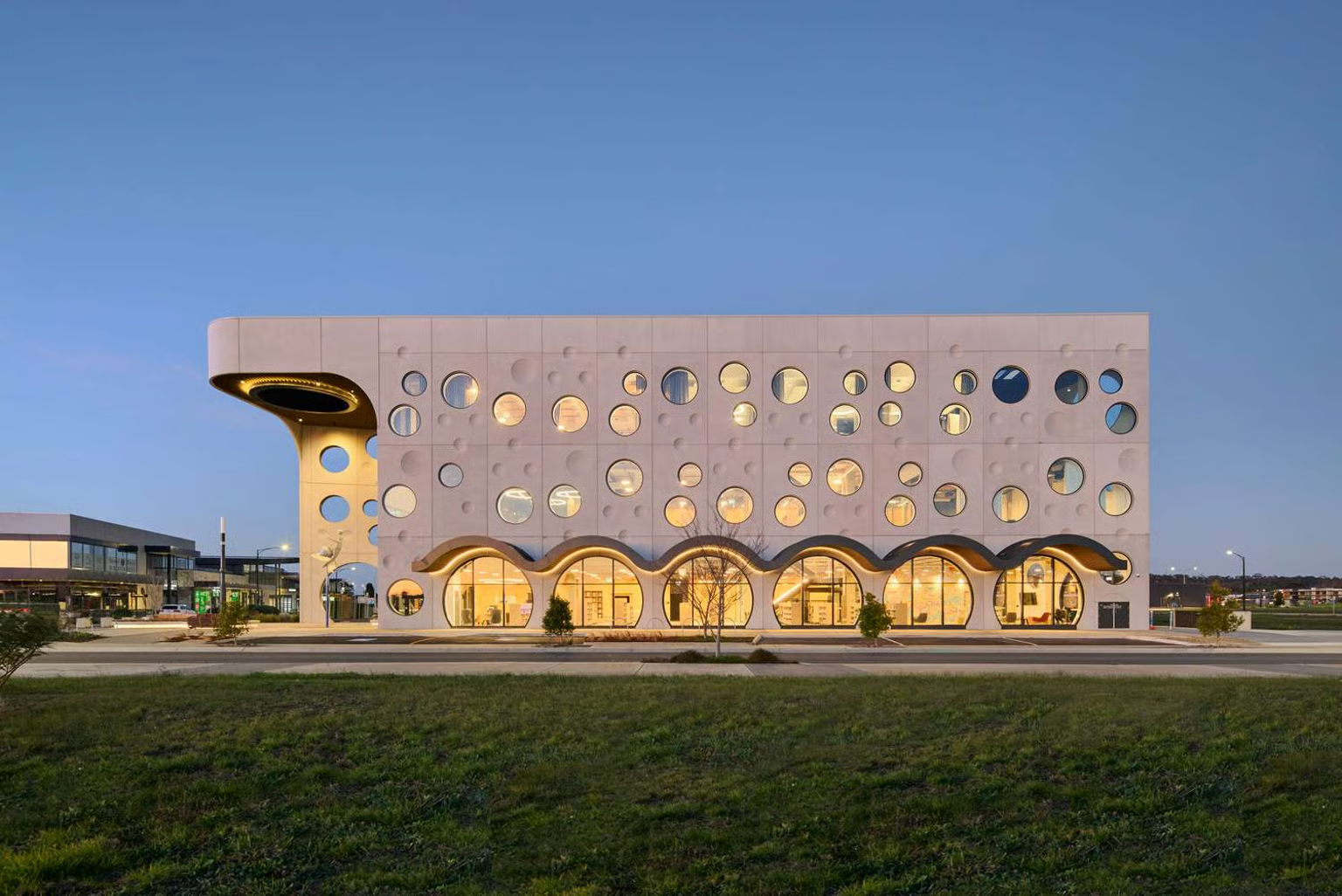 Biyal-a Armstrong Creek Town Centre Library and Community Hub by Buchan. Photography by Tom Roe. Distinctive contemporary building facade featuring a grid of circular windows and sculptural arched forms at ground level, set against a twilight sky in a suburban context.