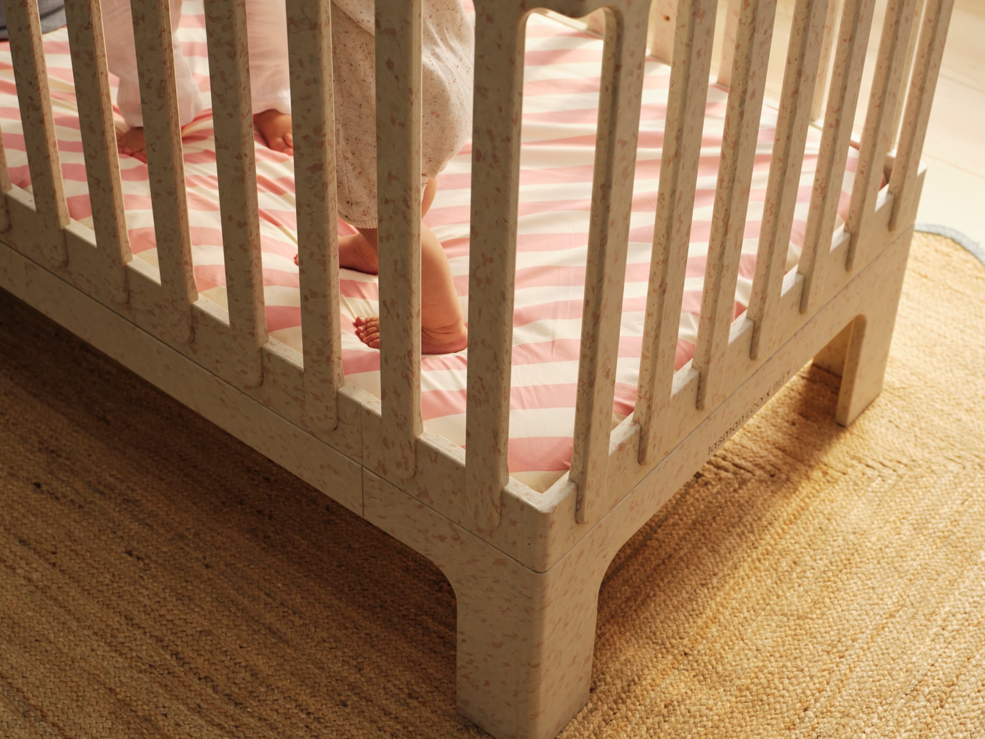Archie Cot by Seena x Defy in Dalmation Pink. Close up of white and pink speckled cot on jute rug with white and pink striped mattress cover. 