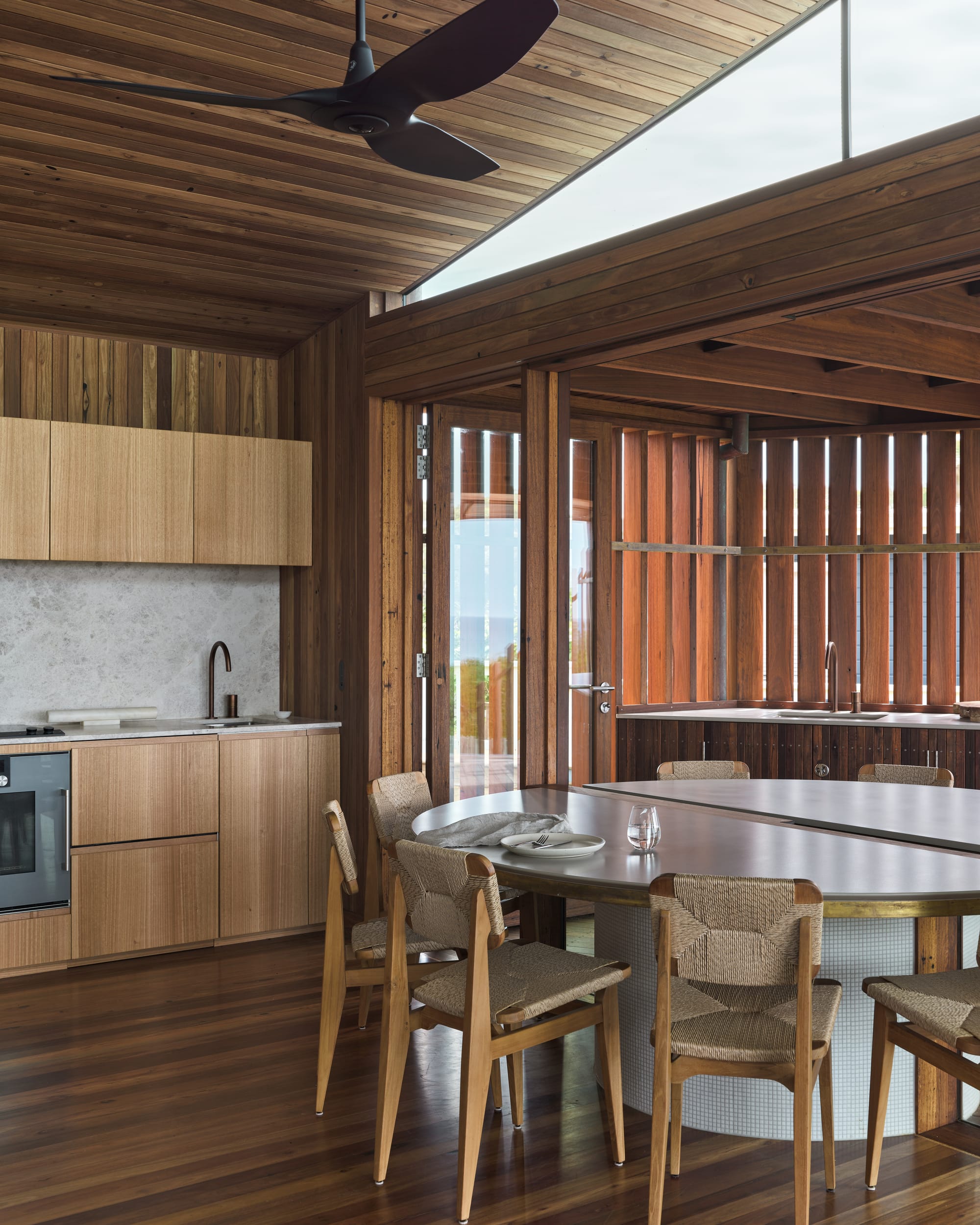 Cake House by Alexander Symes Architect. Photography by Barton Taylor.  Kitchen and dining space with timber walls, floors, ceiling and cabinetry, and a white round dining table with rattan chairs. 