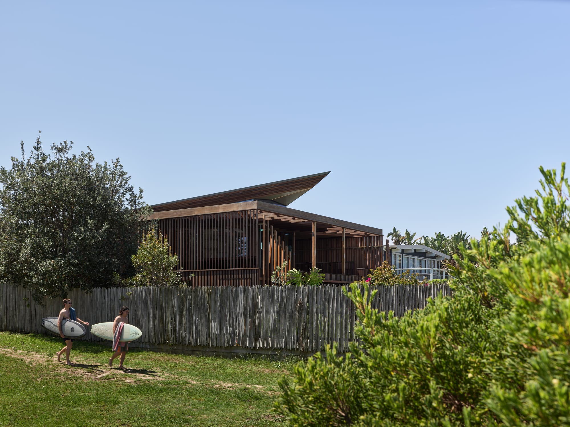 Cake House by Alexander Symes Architect. Photography by Barton Taylor.  Two people with surfboards walk down a sandy path cutting across green grass, in front of a timber fence and timber clad home.