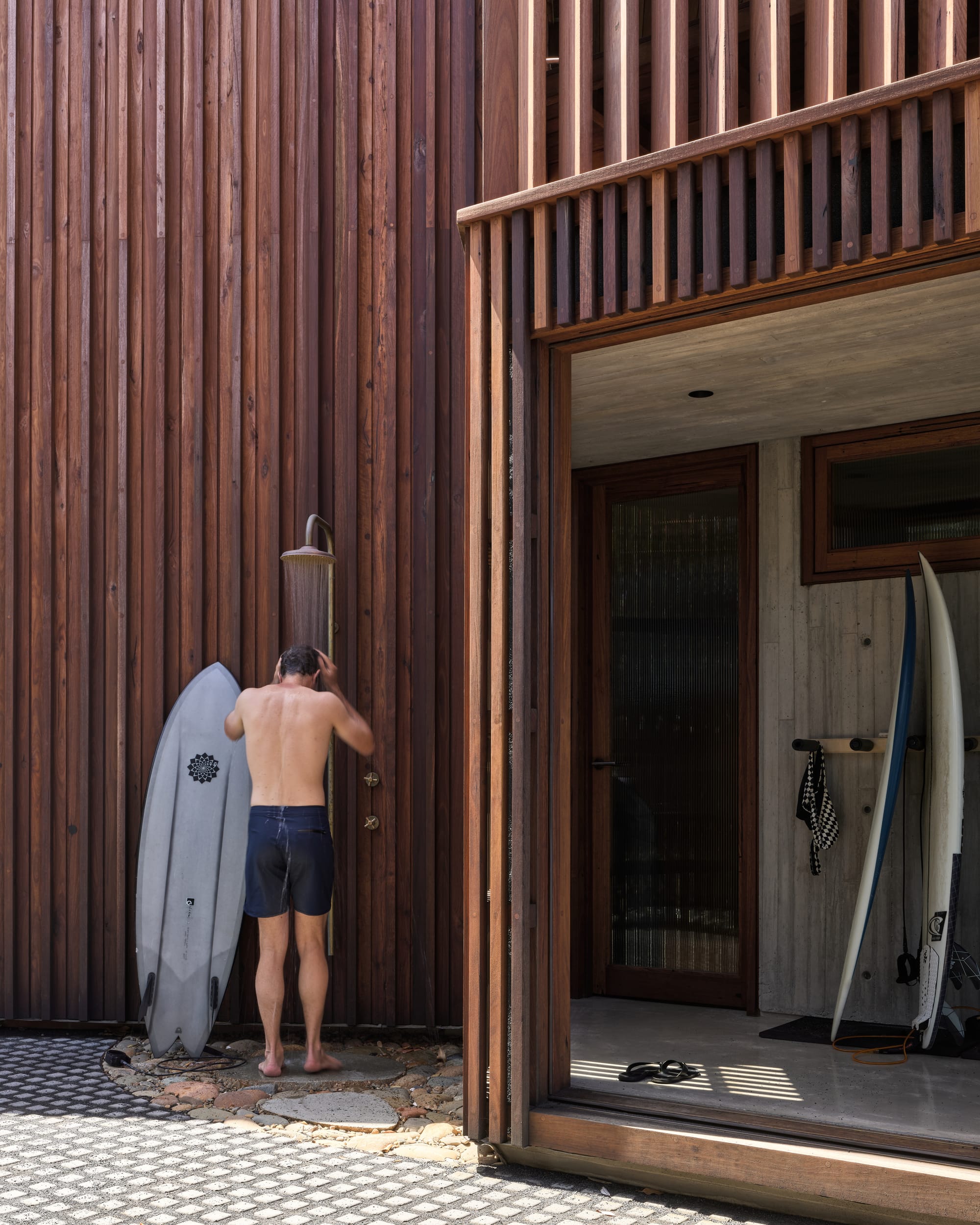 Cake House by Alexander Symes Architect. Photography by Barton Taylor. Man stands next to a surfboard under an outdoor shower in front of a double story timber clad home. 