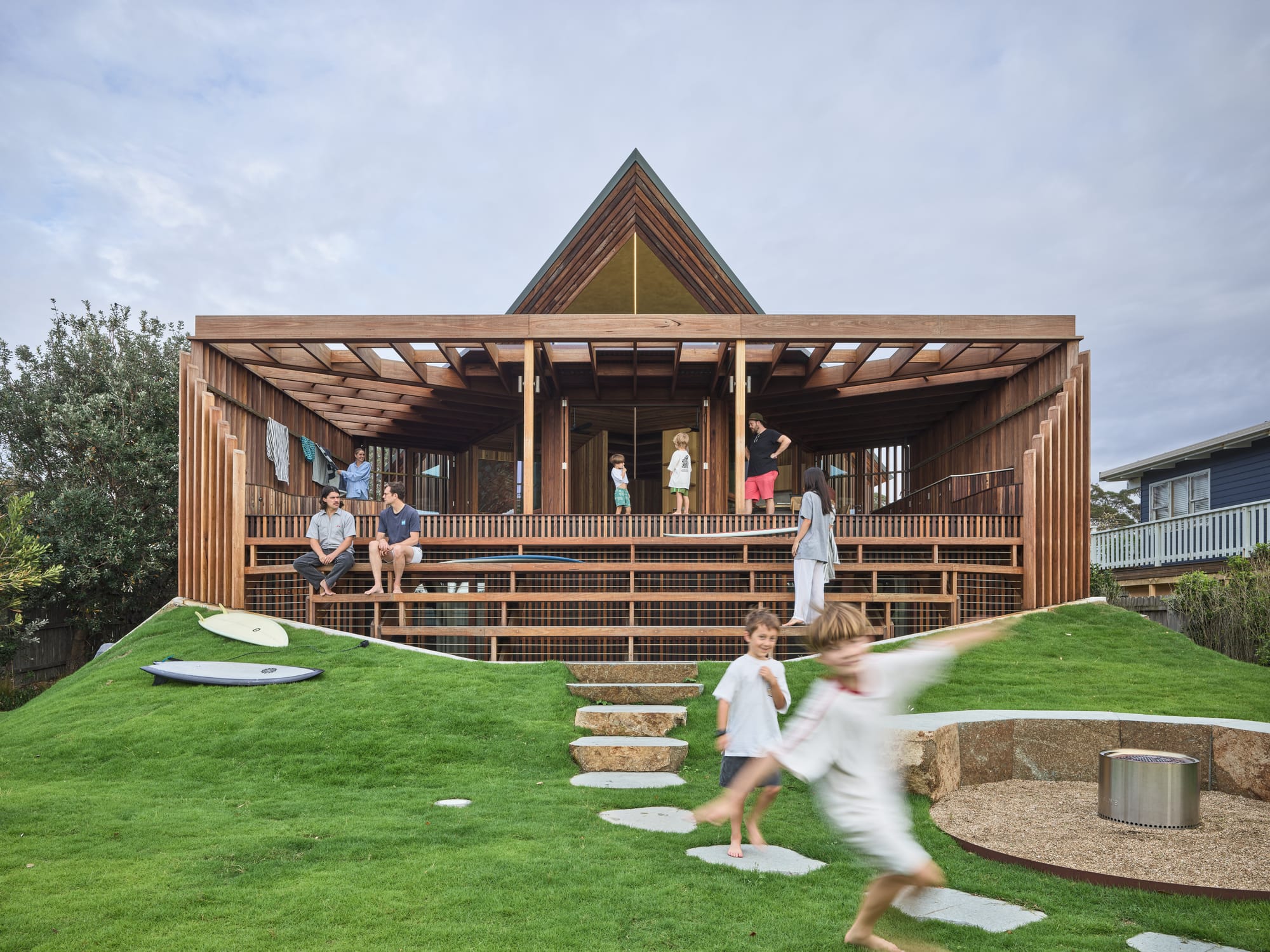 Cake House by Alexander Symes Architect. Photography by Barton Taylor. Man stands next to a surfboard under an outdoor shower in front of a double story timber clad home. 