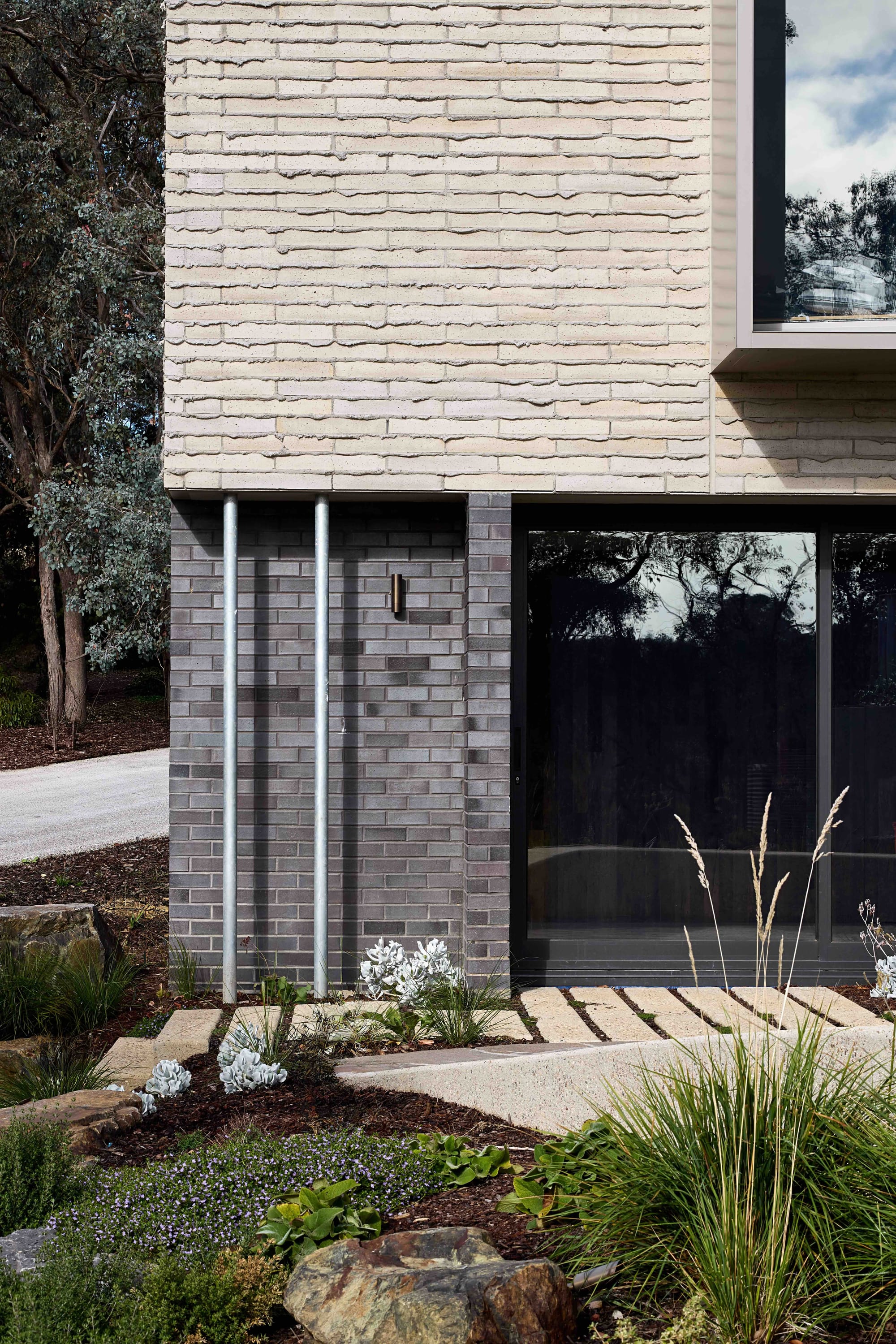 Korinda House by BENT Architecture. Photography by Tatjana Plitt. Close up of exterior of home with dark brick ground floor and timbercrete second story, surrounded by native plants. 