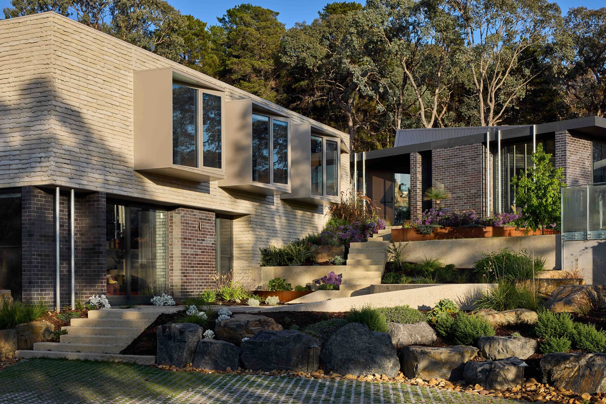 Korinda House by BENT Architecture. Photography by Tatjana Plitt. Exterior of home with tiered stairs and gardens, leading to two story home with dark brick and timbercrete exterior. 
