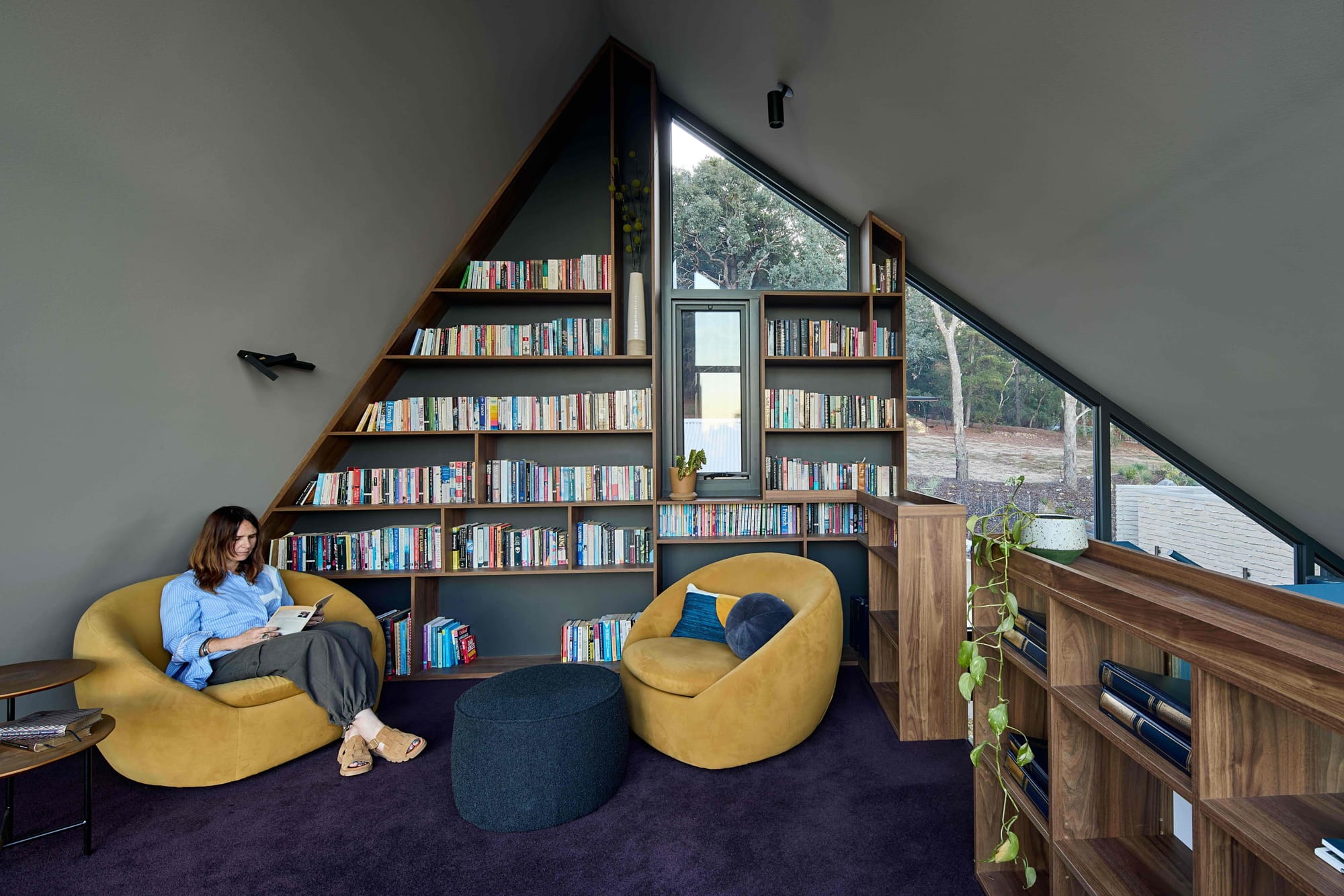 Korinda House by BENT Architecture. Photography by Tatjana Plitt. Library with triangular bookshelf made to fit pitch of roof, and woman sitting on a yellow chair. 