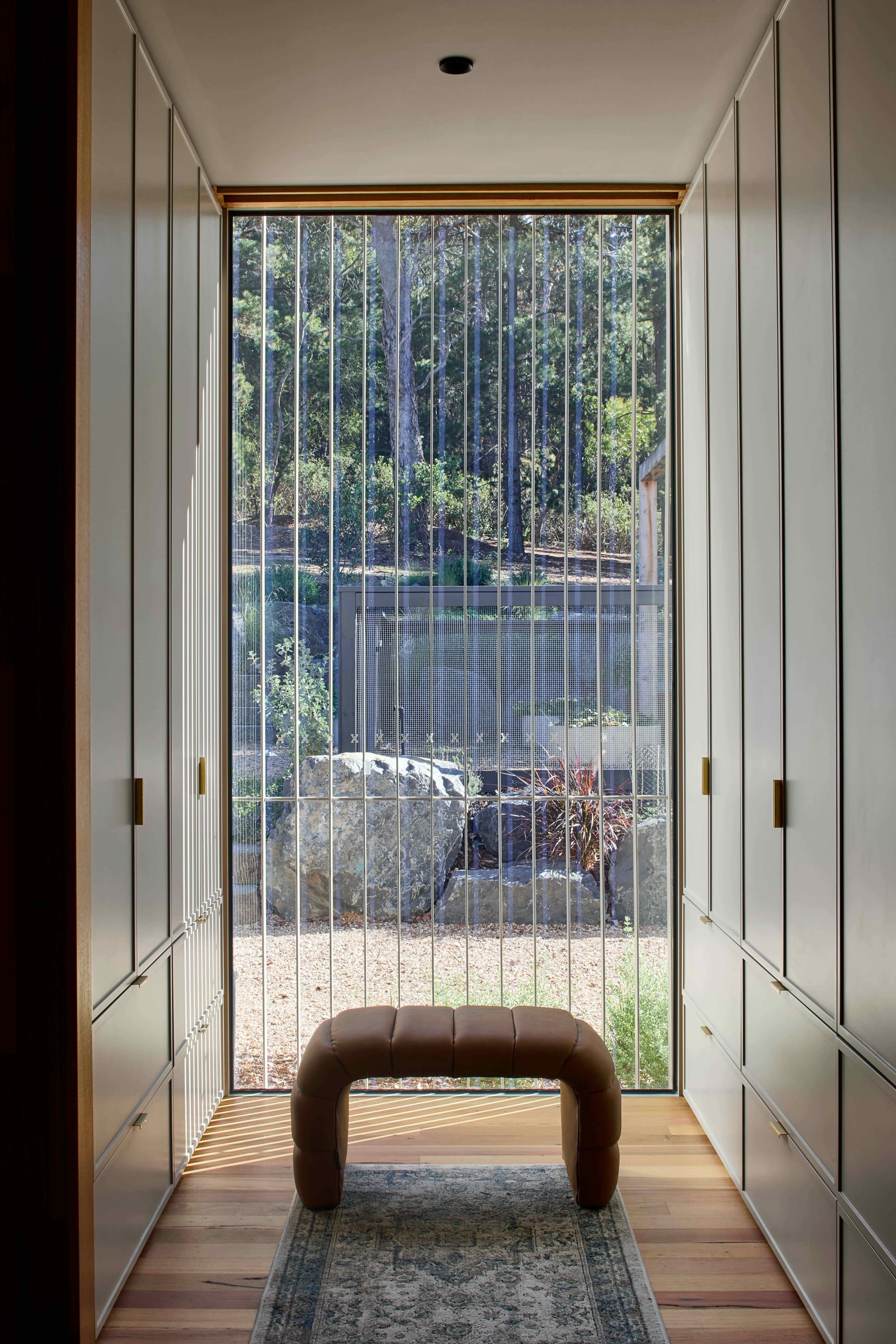 Korinda House by BENT Architecture. Photography by Tatjana Plitt. Walk-in-wardrobe with cabinetry on left and right walls, with glass windows at rear wall with leather stool in front. 