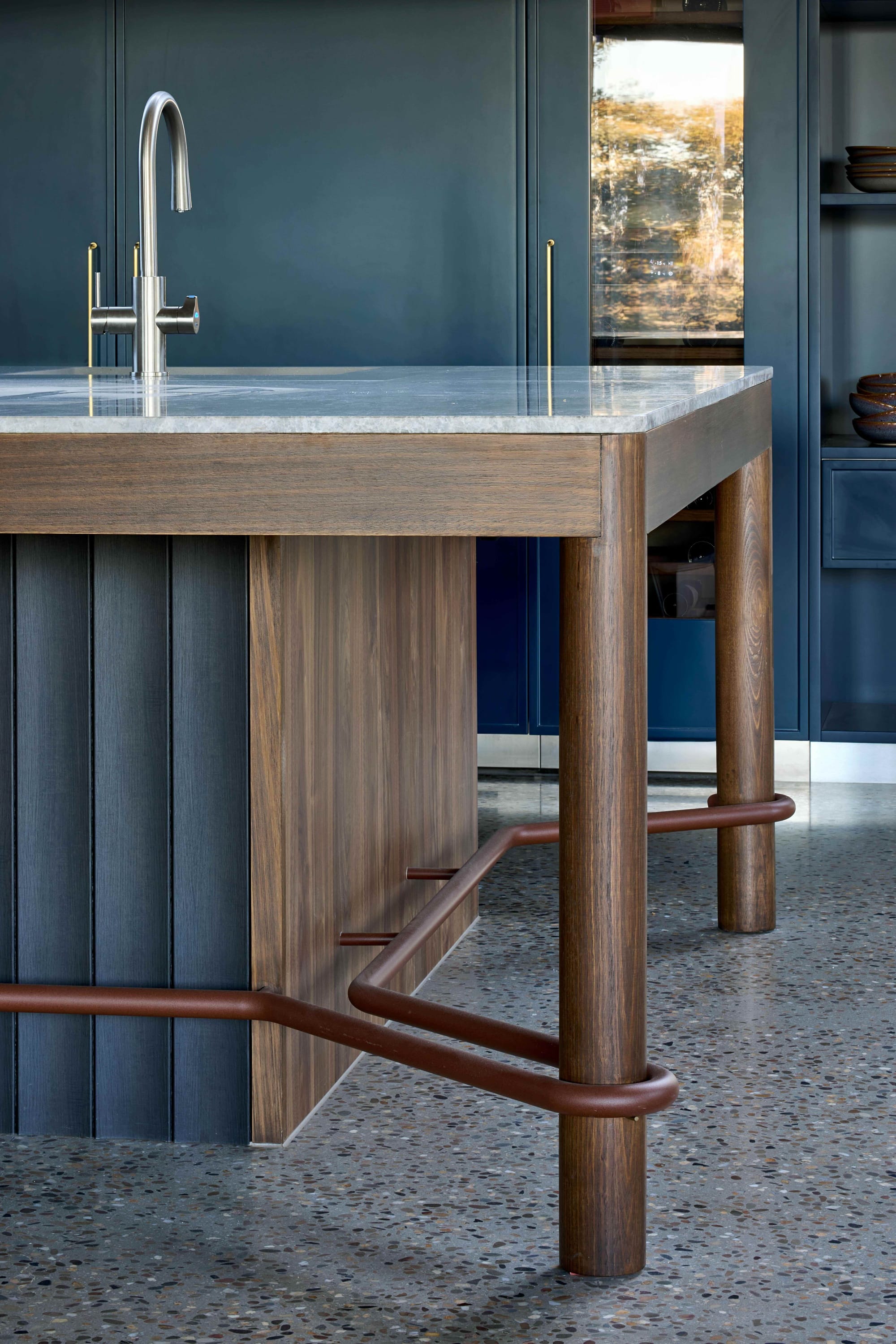 Korinda House by BENT Architecture. Photography by Tatjana Plitt. Detail of kitchen island bench with timber legs, grey kickboard and brown tube footrest wrapping around island and its legs. 