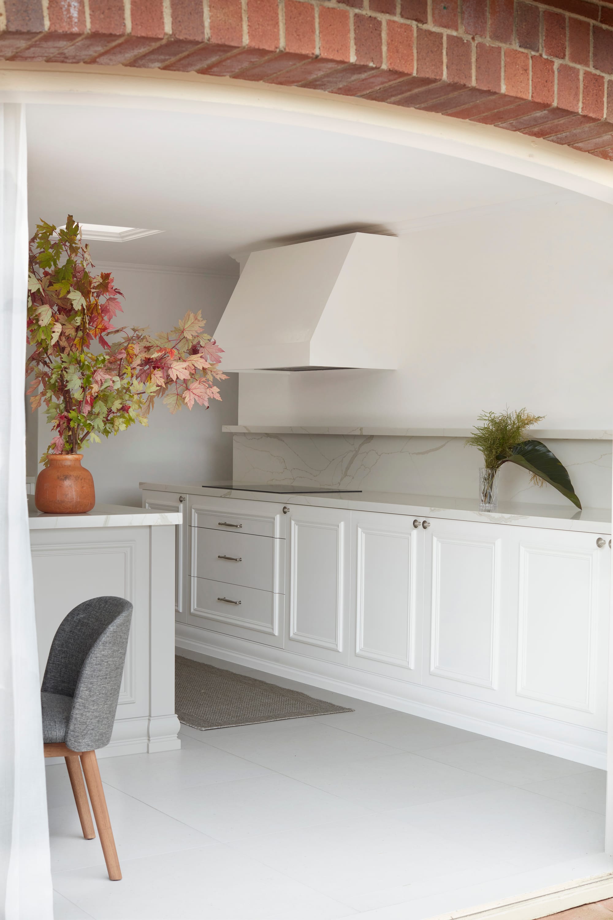 Marathon Road by Marylou Sobel Interior Design. Photography by Prue Ruscoe. Kitchen with white tile flooring, white cabinetry, marble splashback and red brick archway connecting inside and out. 