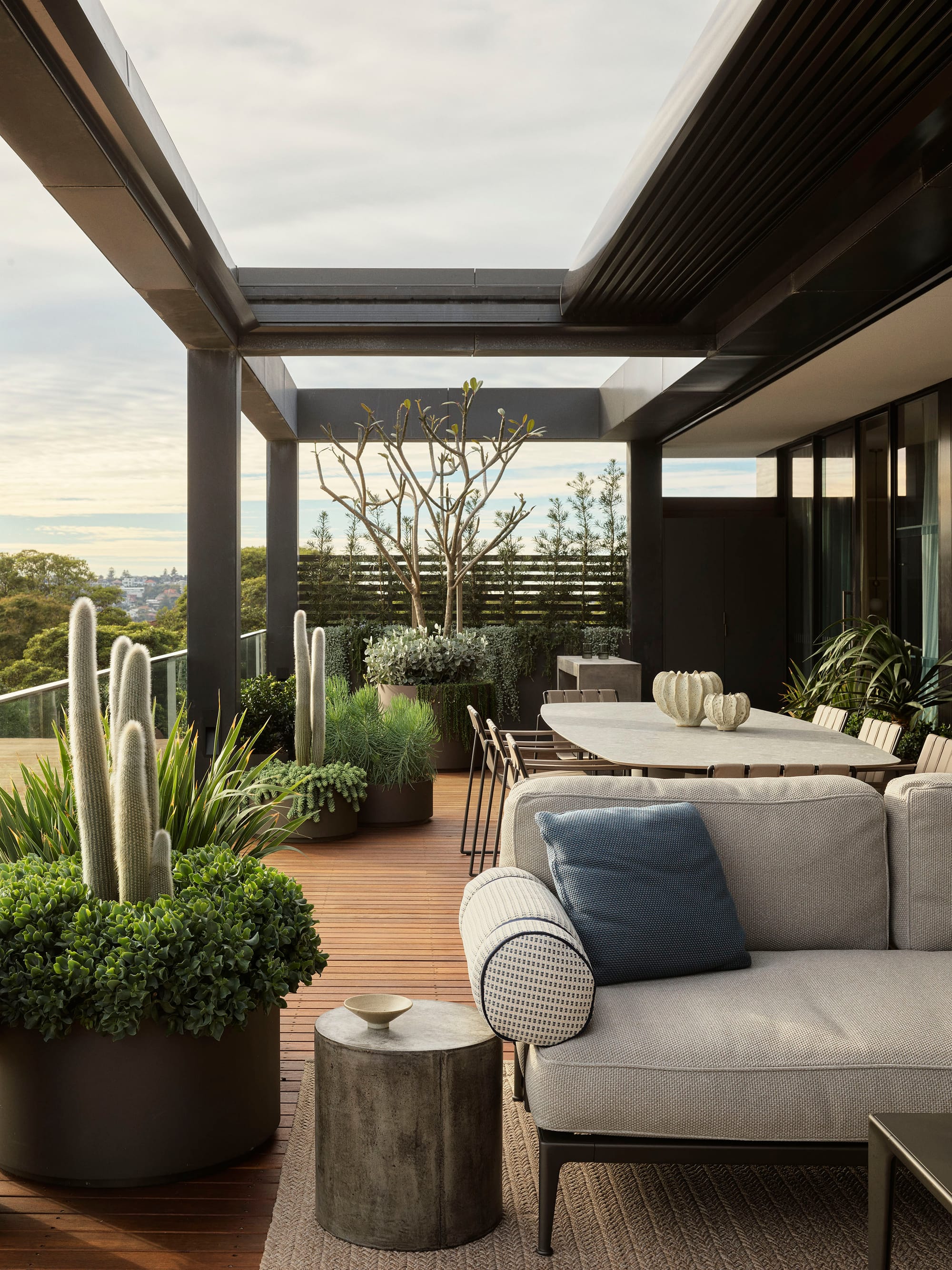 Bundarra Penthouse by Marylou Sobel Interior Design. Photography by Anson Smart. Rooftop patio with grey outdoor furniture, timber decking and large plant pots around glass balustrade. 