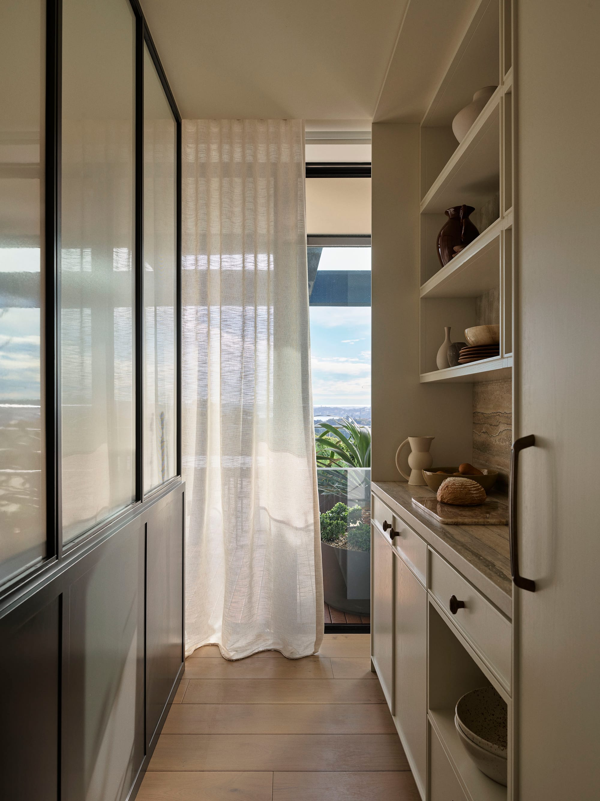 Bundarra Penthouse by Marylou Sobel Interior Design. Photography by Anson Smart. Scullery kitchen with integrated white open-face cabinetry on right side, black cabinetry with frosted glass on left and full height windows at end with landscape views. 