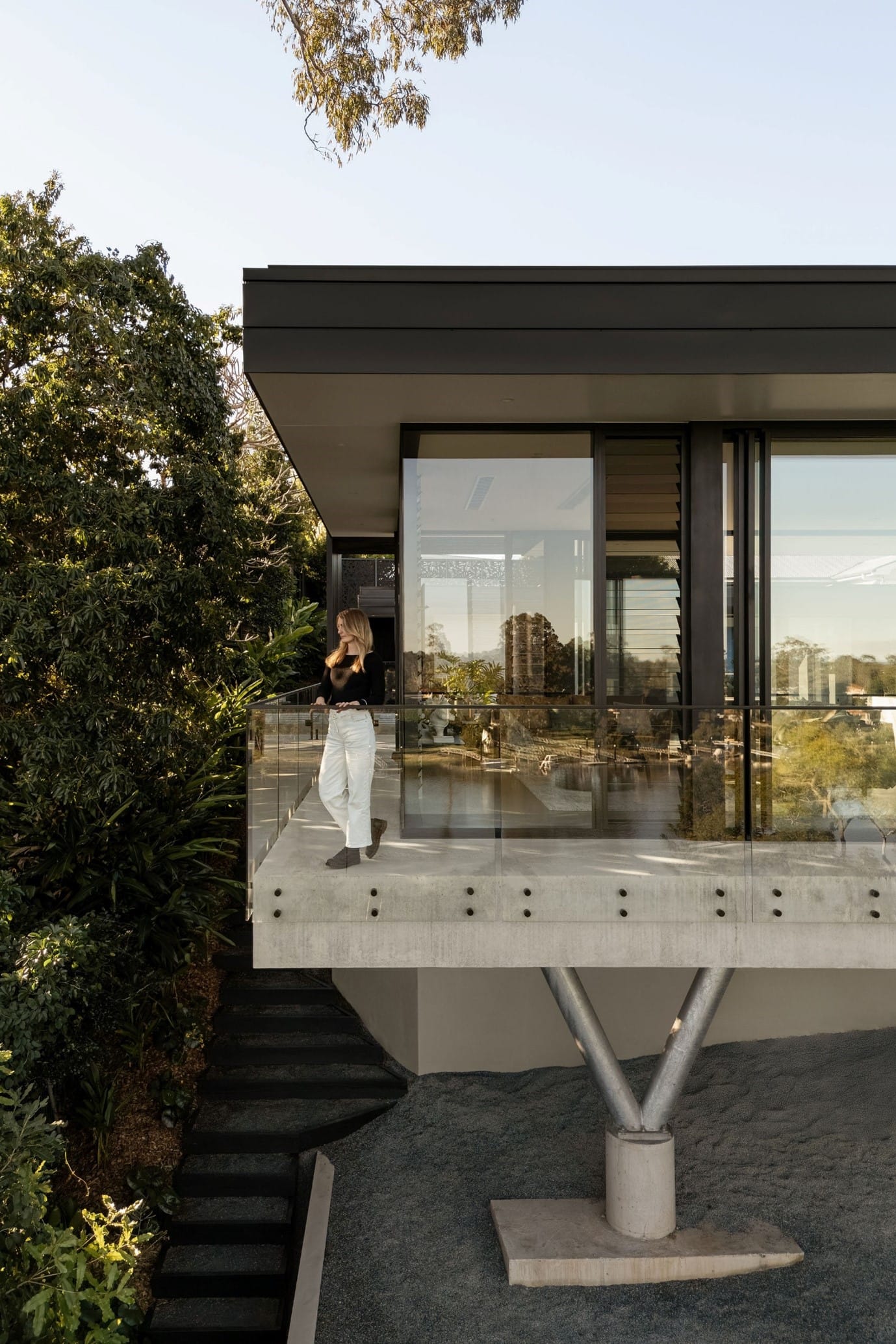 The River House, Indooroopilly by Sarah Waller Architecture. Photography by Alyne Media. Woman stands at the corner of a balcony, behind a glass balustrade in front of residential home with concrete floors and glass windows and doors. 