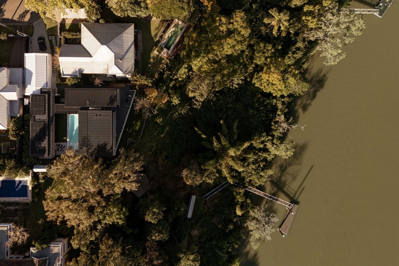 The River House, Indooroopilly by Sarah Waller Architecture. Photography by Alyne Media. Aerial view of large river, with dense foliage on river banks, and a black metal roof of a residential home to the left. 