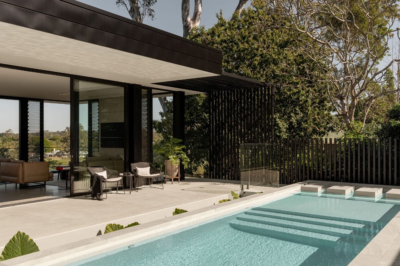 The River House, Indooroopilly by Sarah Waller Architecture. Photography by Alyne Media. Rear patio of home with lap pool, concrete flooring, and glass windows opening onto interior living area. 