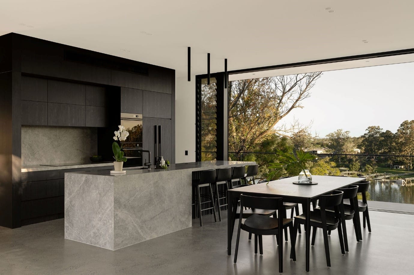The River House, Indooroopilly by Sarah Waller Architecture. Photography by Alyne Media. Kitchen with black cabinetry, grey stone island bench, black stools and dining set. 