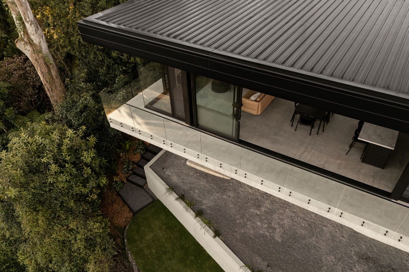 The River House, Indooroopilly by Sarah Waller Architecture. Photography by Alyne Media. High angle shot of angled balcony with poured concrete floors, glass balustrade and flat, black tin roof. 