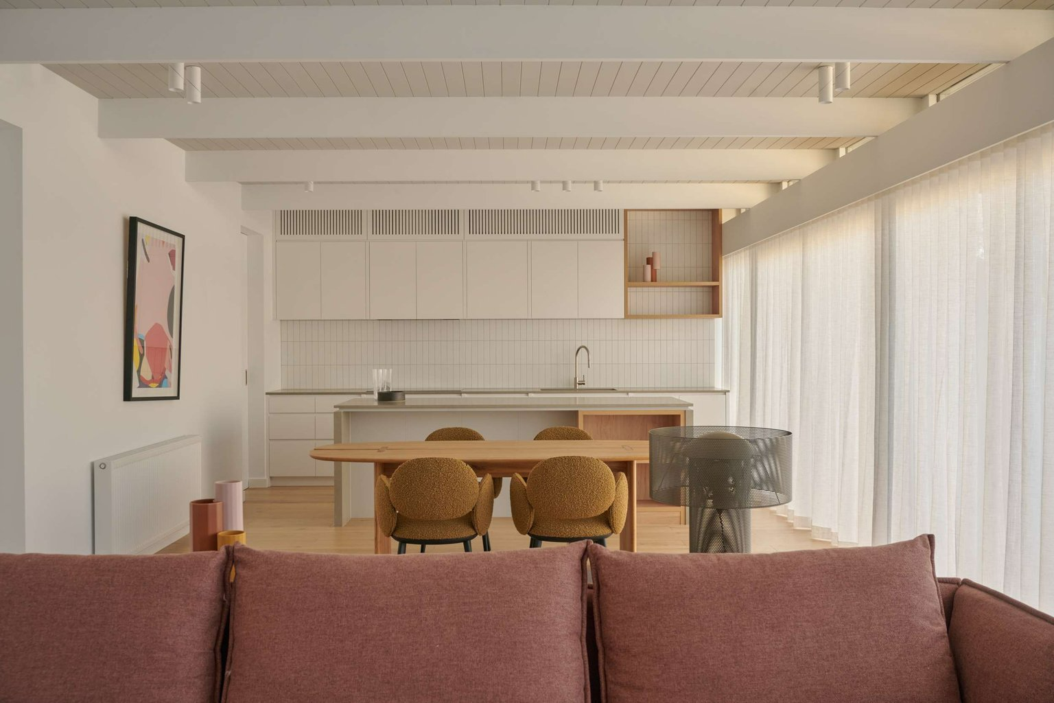 Moonya Modern by FYC Architects. Photography by Veeral Patel. Open plan kitchen, living and dining room with exposed ceiling beams, white kitchen, timber dining table and pink sofa. 