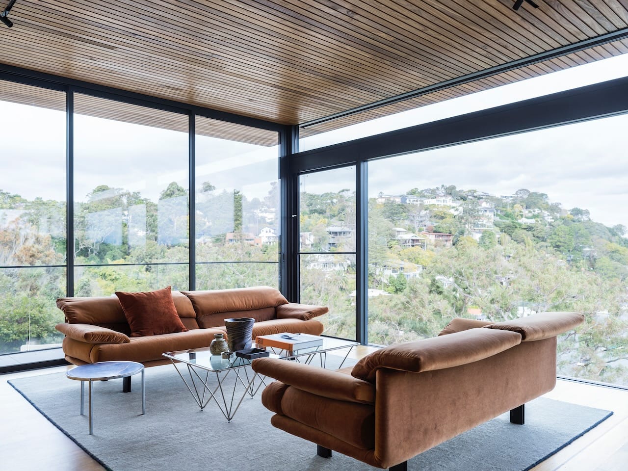 Corbel House by Nick Bell Architects. Photography by Justin Alexander. Living space with two brown couches and full height windows on all walls with panoramic views over bushland.