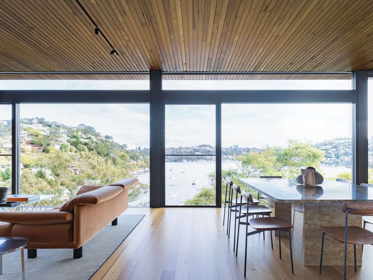 Corbel House by Nick Bell Architects. Photography by Justin Alexander. Open plan dining and living space with timber floors, brown couch, granite dining table and full height windows overlooking harbour and hills. 