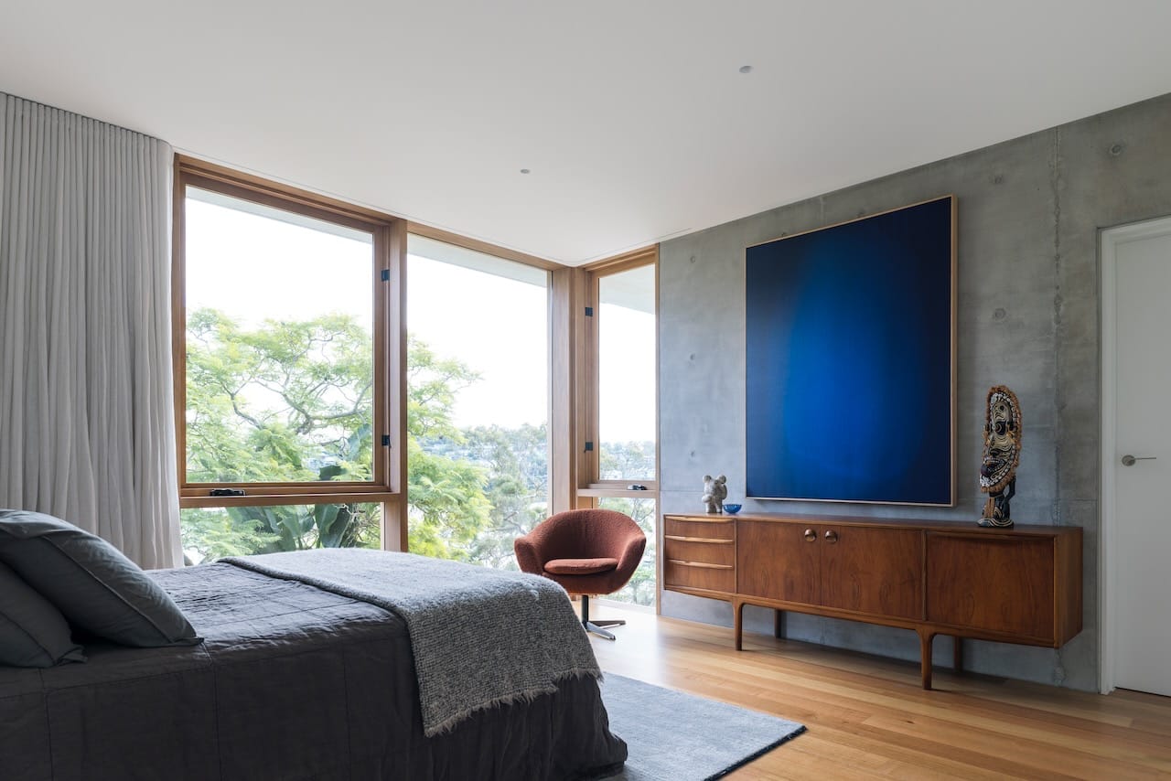 Corbel House by Nick Bell Architects. Photography by Justin Alexander. Bedroom with concrete walls, electric blue artwork, a timber sideboard and full height windows overlooking tall tree. 