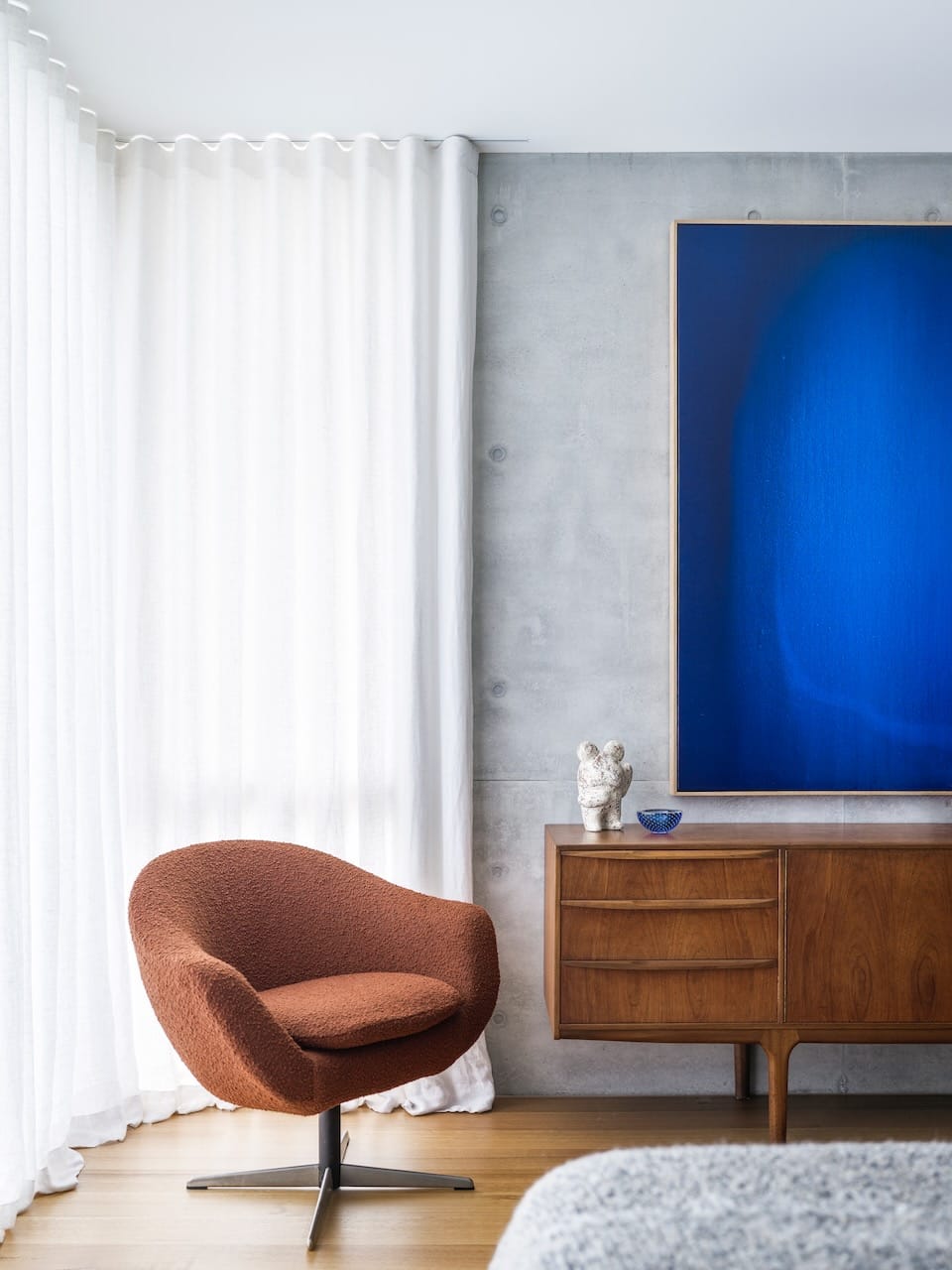 Corbel House by Nick Bell Architects. Photography by Justin Alexander. Terracotta boucle armchair in front of white sheer curtains, next to MCM sideboard and electric blue artwork. 